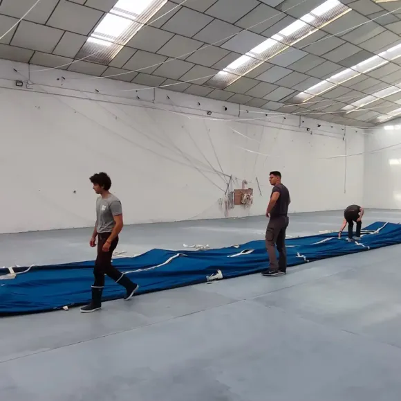 Personas en un almacén doblando una sábana azul larga. Algunos caminan, otros trabajan con ella. Paredes blancas.