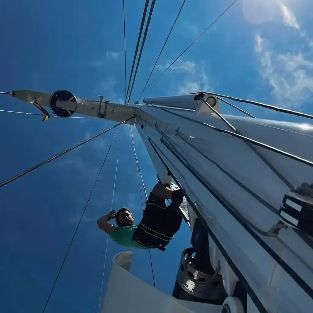 Hombre trepando al mástil de un velero contra un cielo azul.