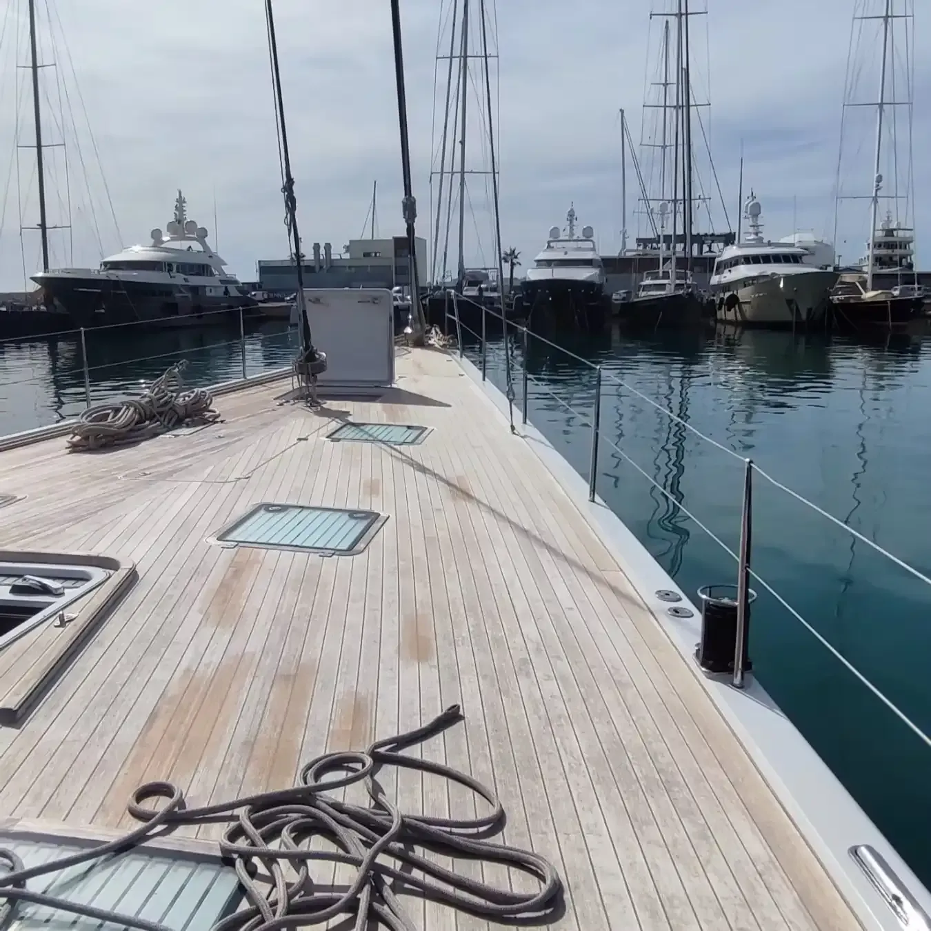 Cubierta de madera de un velero en un puerto, otros barcos al fondo en un día soleado.