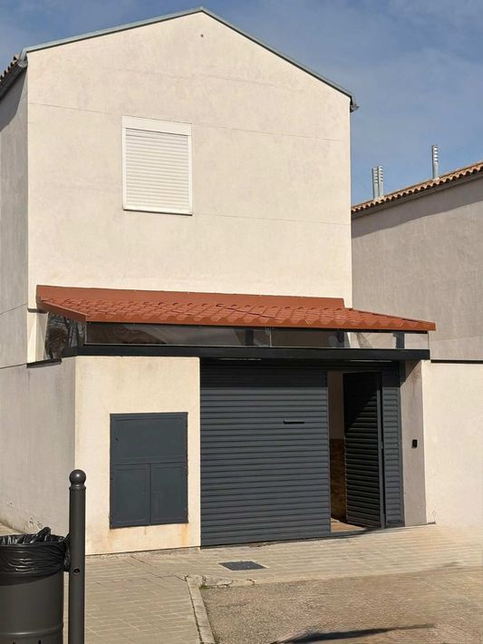 Pequeño edificio blanco con toldo rojo y puerta de garaje gris en una calle empedrada.