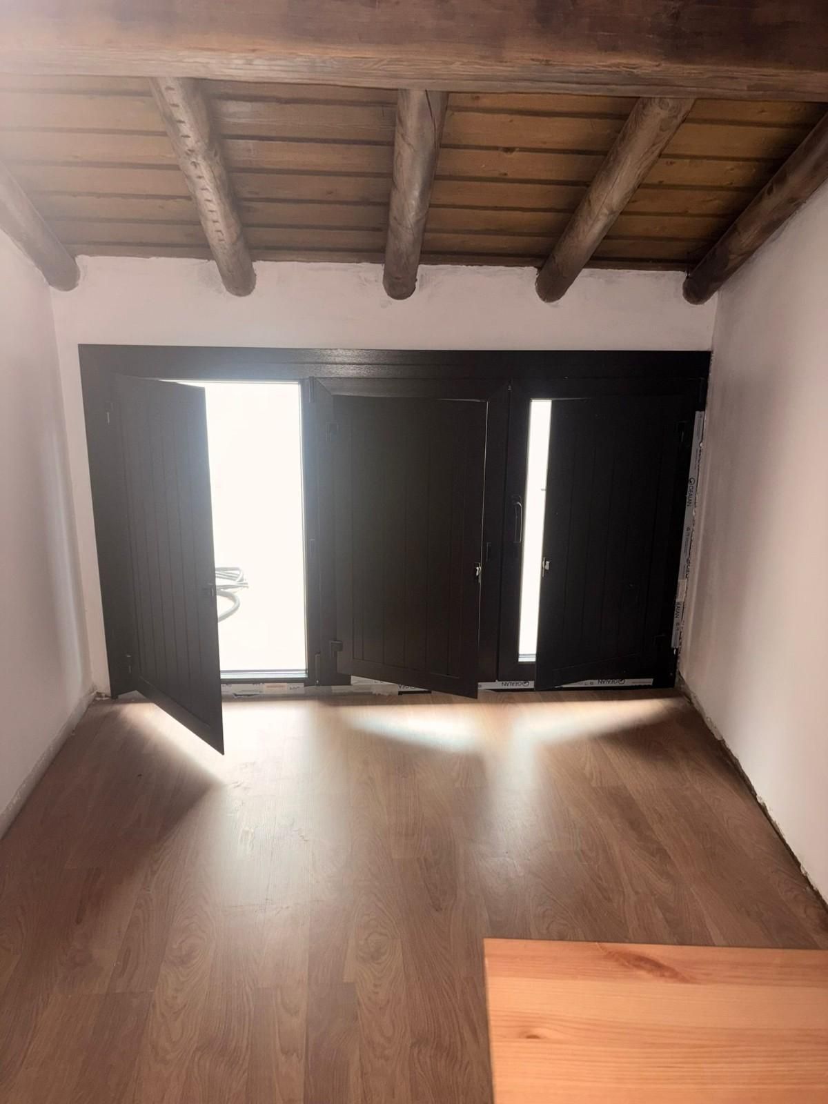 Habitación vacía con suelo de madera, techo con vigas vistas y puertas dobles negras abiertas que dejan entrar mucha luz.
