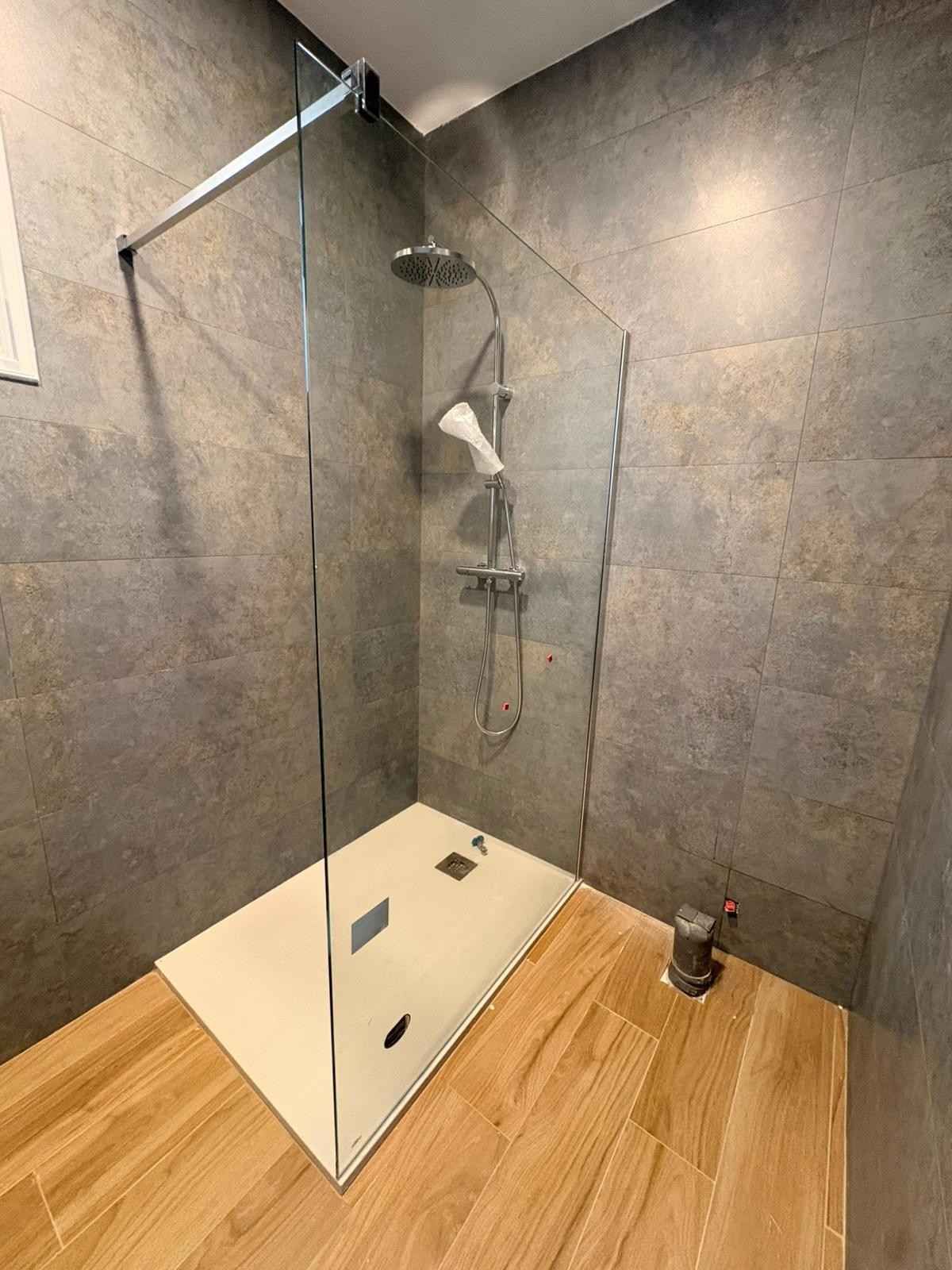 Moderna ducha a ras de suelo con mampara de cristal, paredes de azulejos grises, base blanca y suelo con aspecto de madera.
