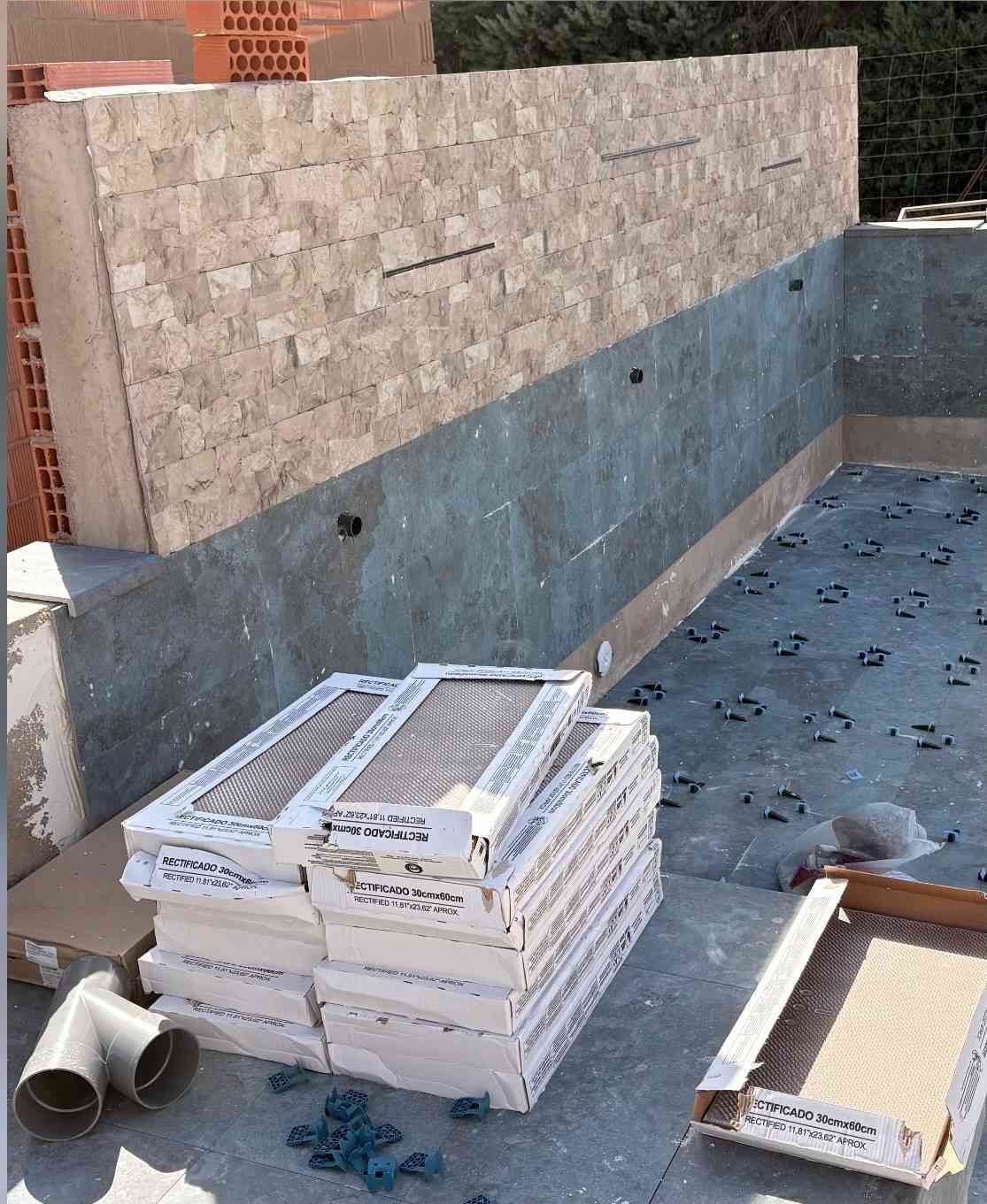 Piscina en construcción con azulejos apilados y una pared parcialmente alicatada junto a una impermeabilización azul.