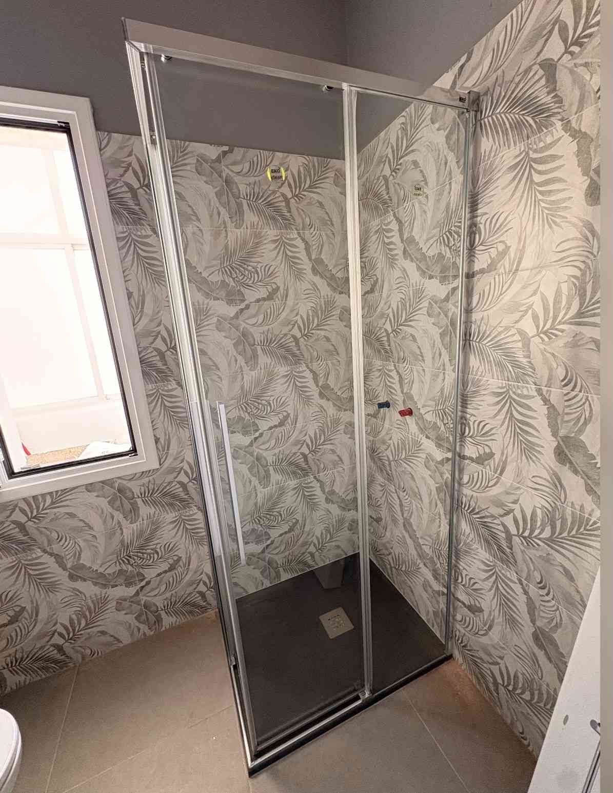 Baño pequeño con cabina de ducha de cristal, papel pintado con estampado de hojas y una ventana.