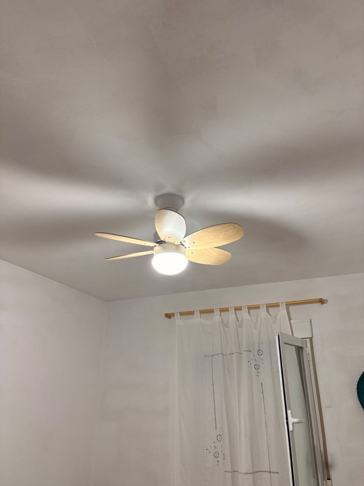 Ventilador de techo con luz y aspas doradas en un dormitorio blanco, sobre una ventana con cortinas y un espejo.