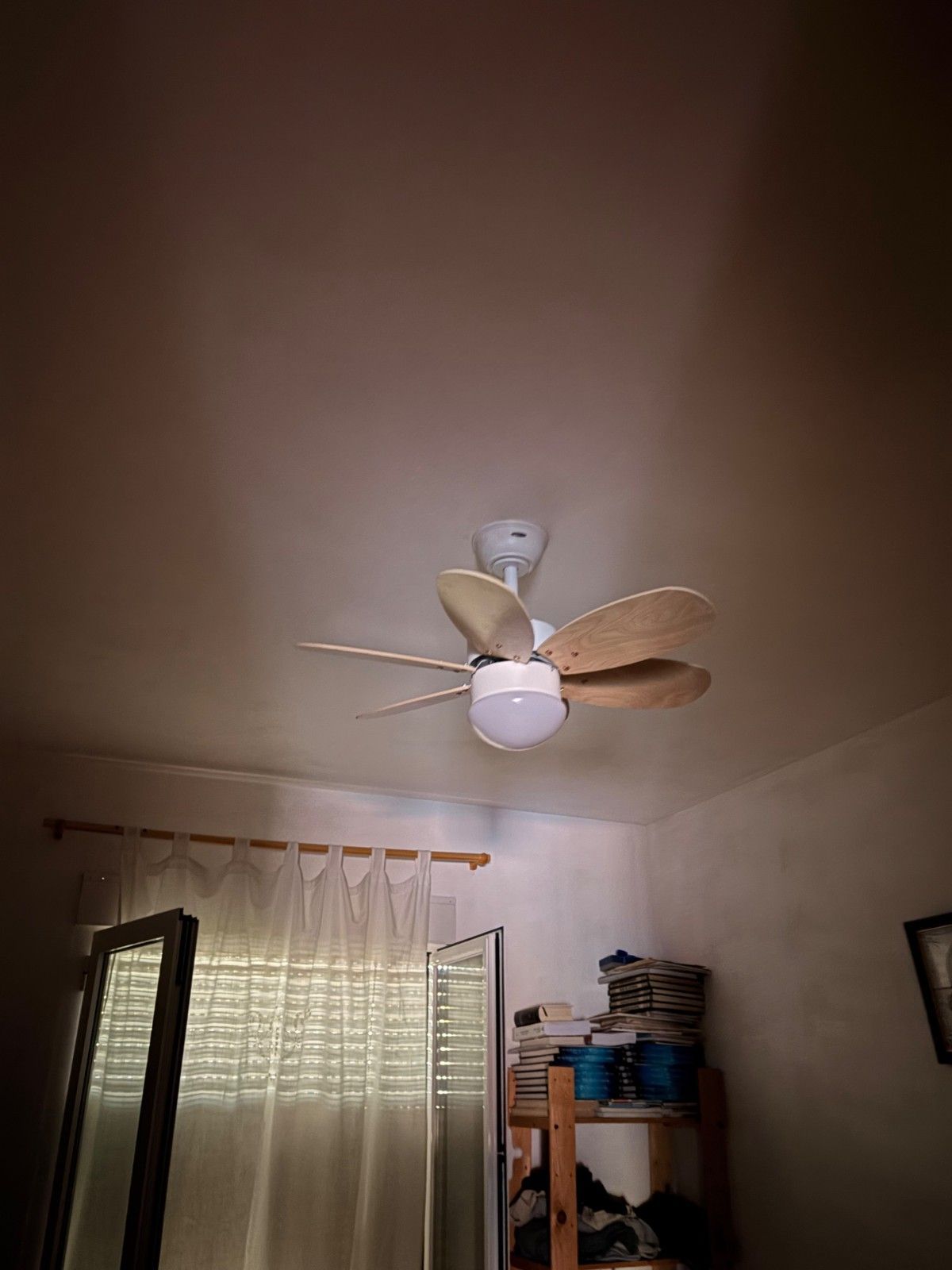 Ventilador de techo y ventana con cortinas en un dormitorio pequeño con estanterías.