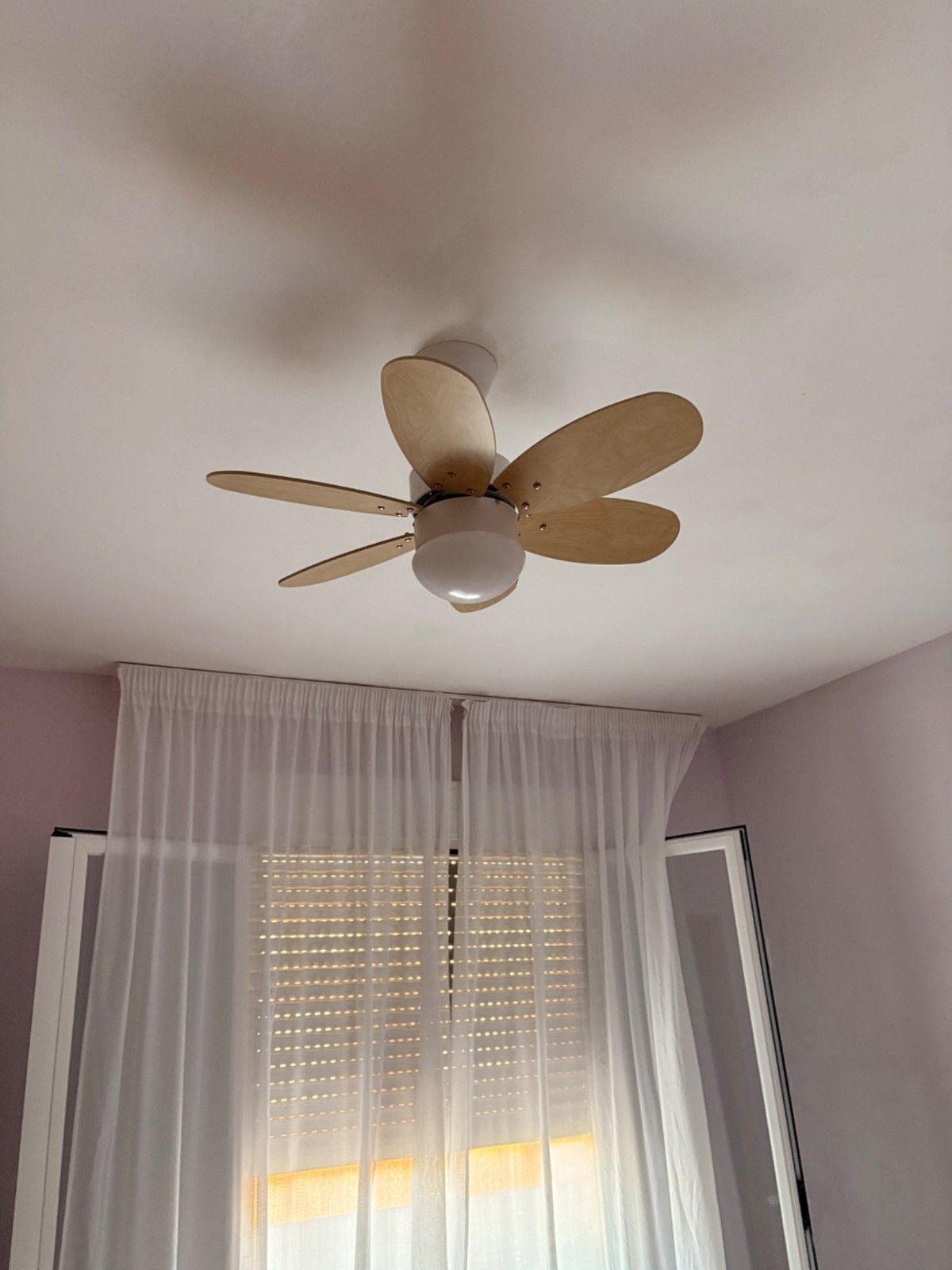 Ventilador de techo sobre una ventana con cortinas transparentes en un dormitorio con iluminación tenue.