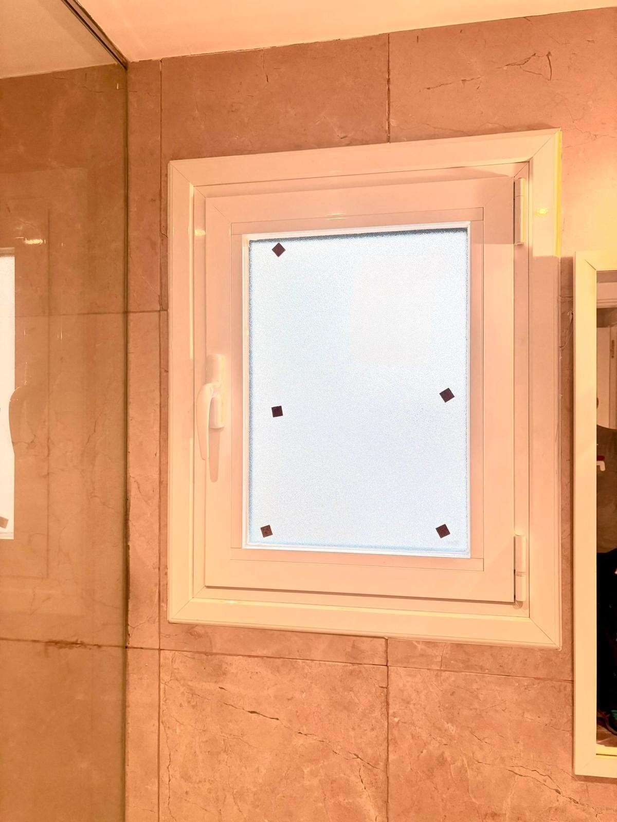 Ventana blanca en el baño, en una pared de azulejos beige, parcialmente abierta a un panel de vidrio esmerilado.