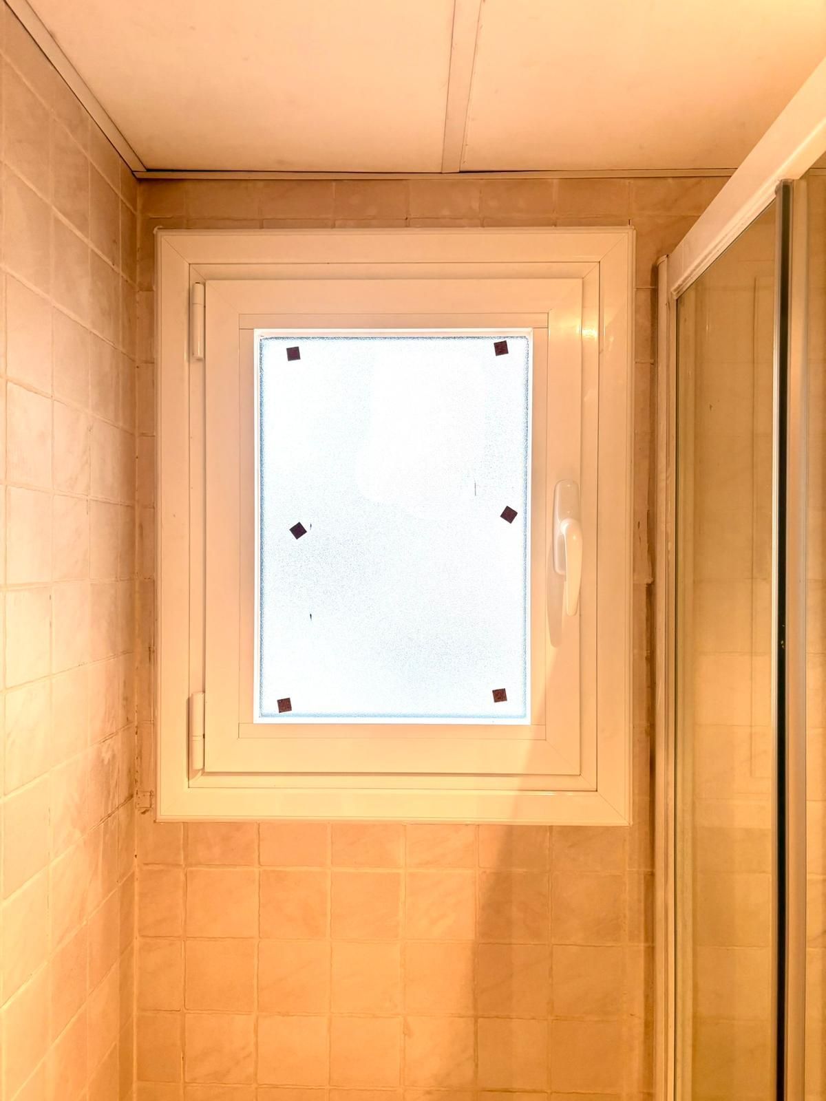 Pequeña ventana esmerilada en una esquina de la pared y el techo de un baño revestido de azulejos beige.