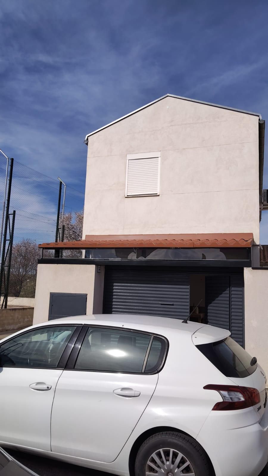 Un hatchback blanco estacionado frente a una pequeña casa beige de dos pisos bajo un cielo azul.