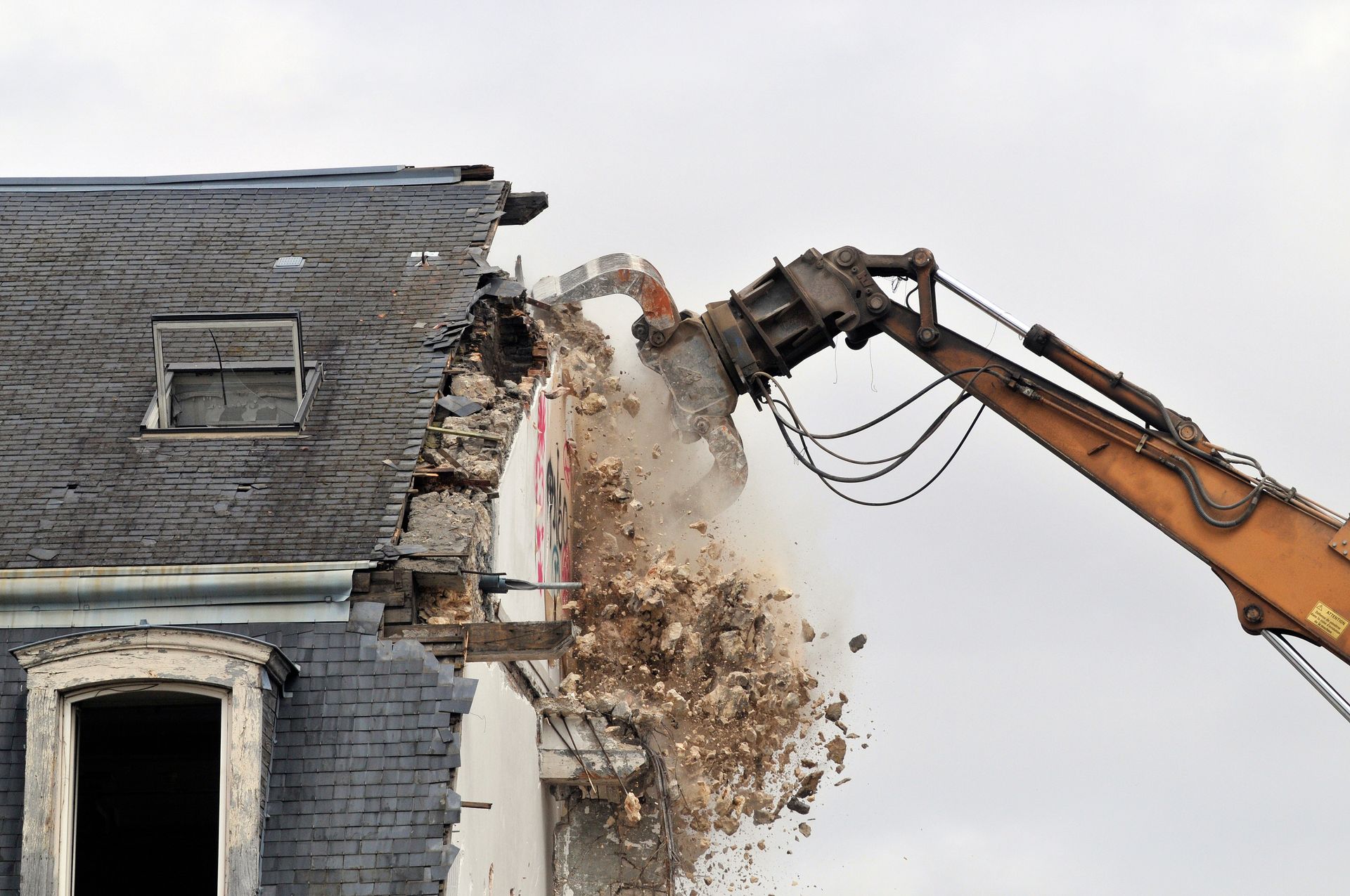 Une pelleteuse démolit une maison en briques, soulevant un nuage de poussière sur un ciel gris.