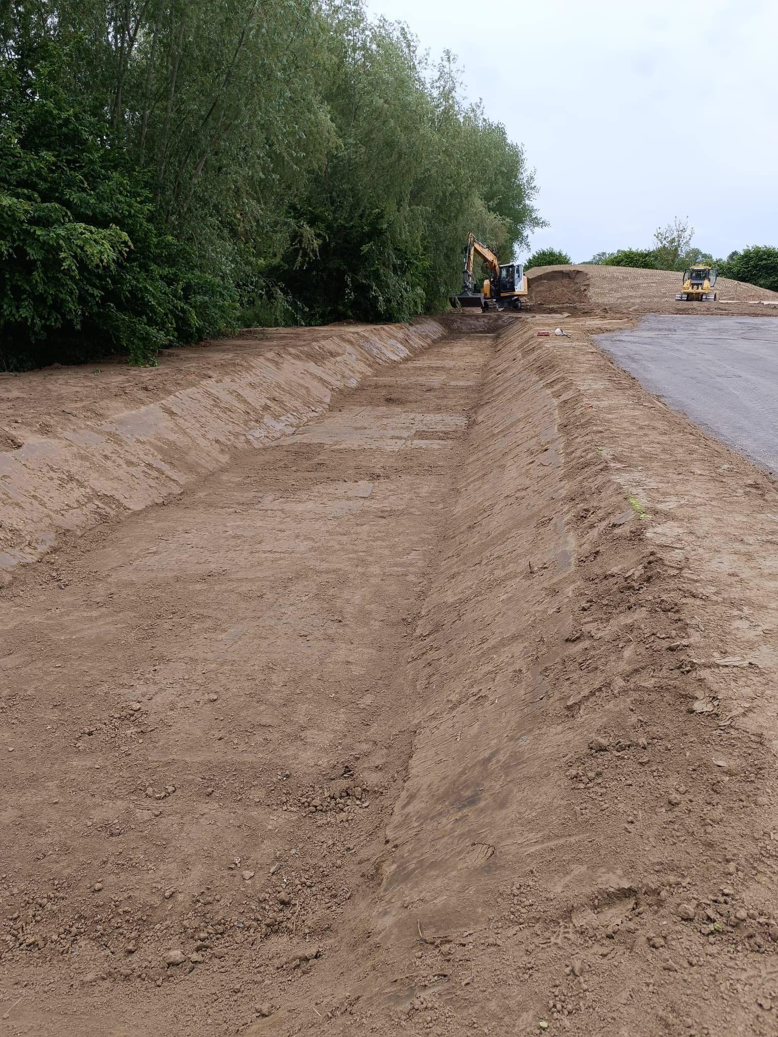 Un fossé creusé le long d'une route, avec une excavatrice en activité à l'arrière-plan et un tas de terre.