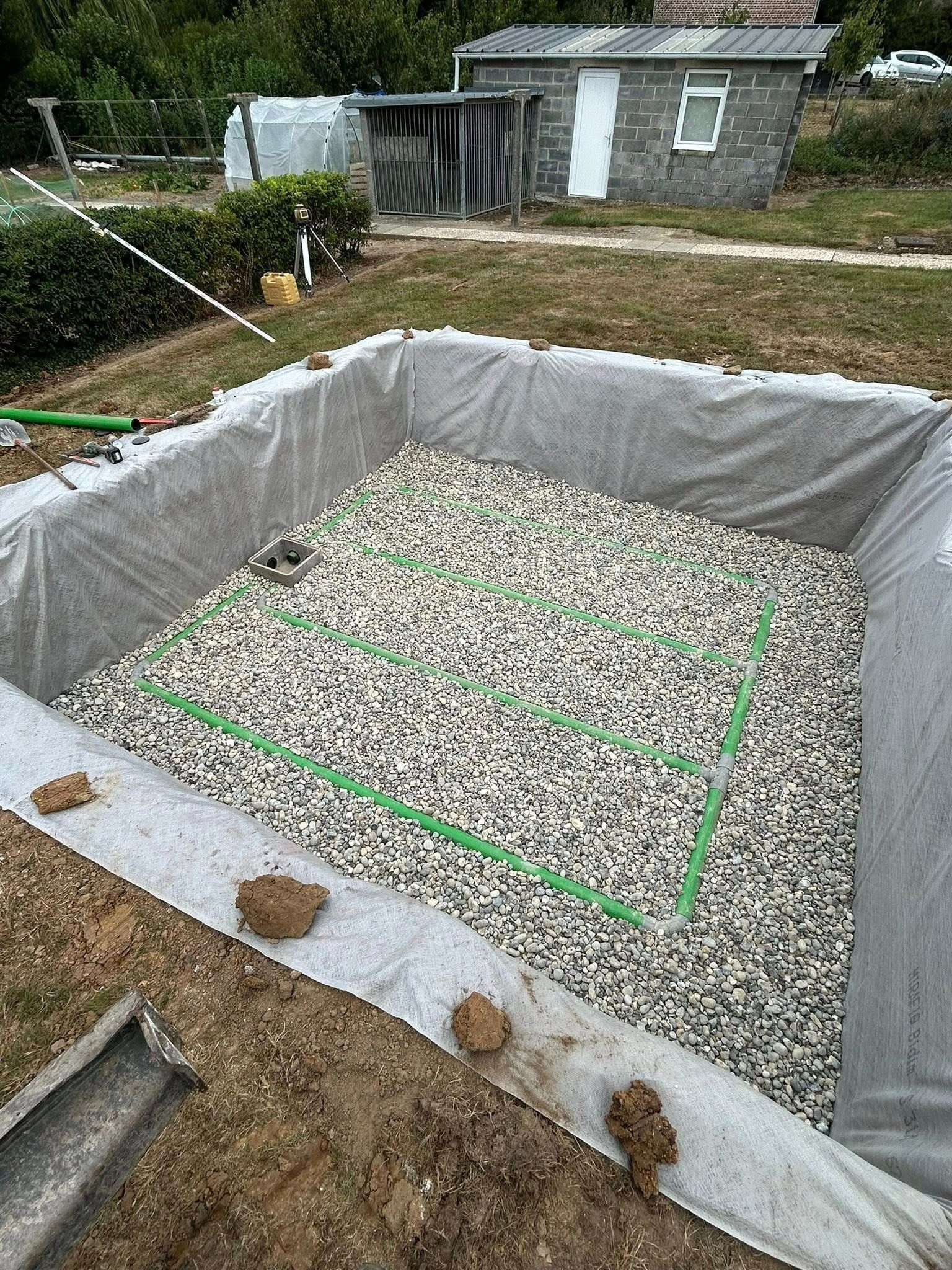 Un bassin de jardin en construction, rempli de gravier, recouvert de tissu gris et entouré d'une bordure de terre.