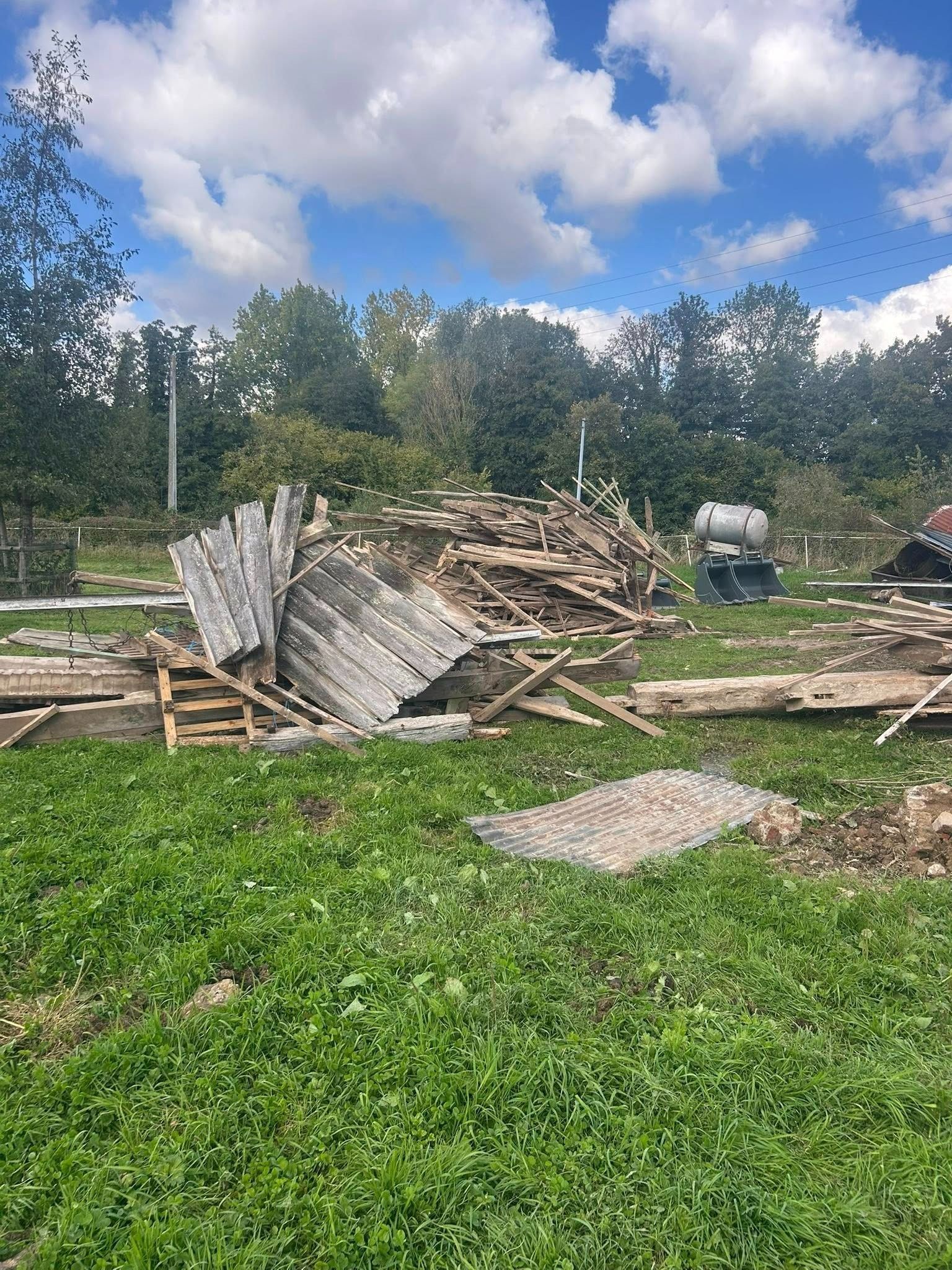Tas de débris de vieux bois et de tôle ondulée sur fond d'herbe verte, d'arbres et de ciel bleu.