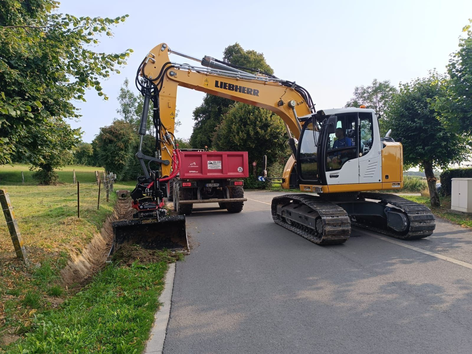Une pelleteuse jaune charge un camion-benne rouge sur une route longeant un fossé herbeux.