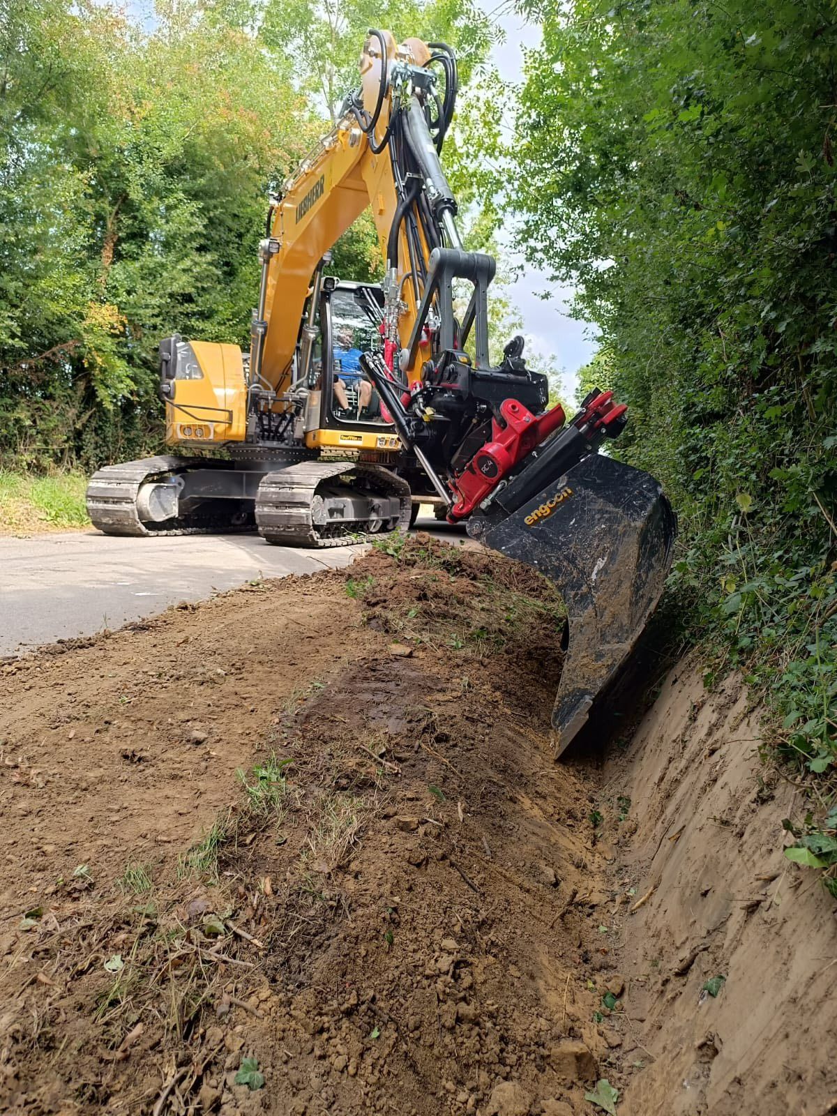 Une pelleteuse jaune creuse un fossé au bord d'une route, avec des arbres en arrière-plan.