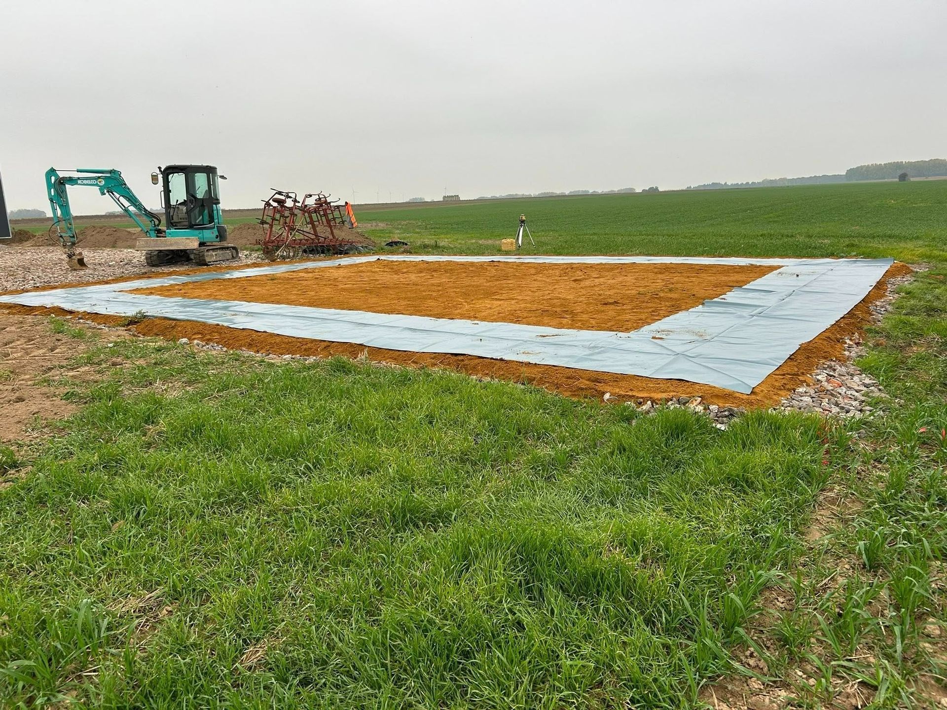 Chantier de construction dans un champ avec une petite excavatrice, délimité par un carré et de l'herbe verte.