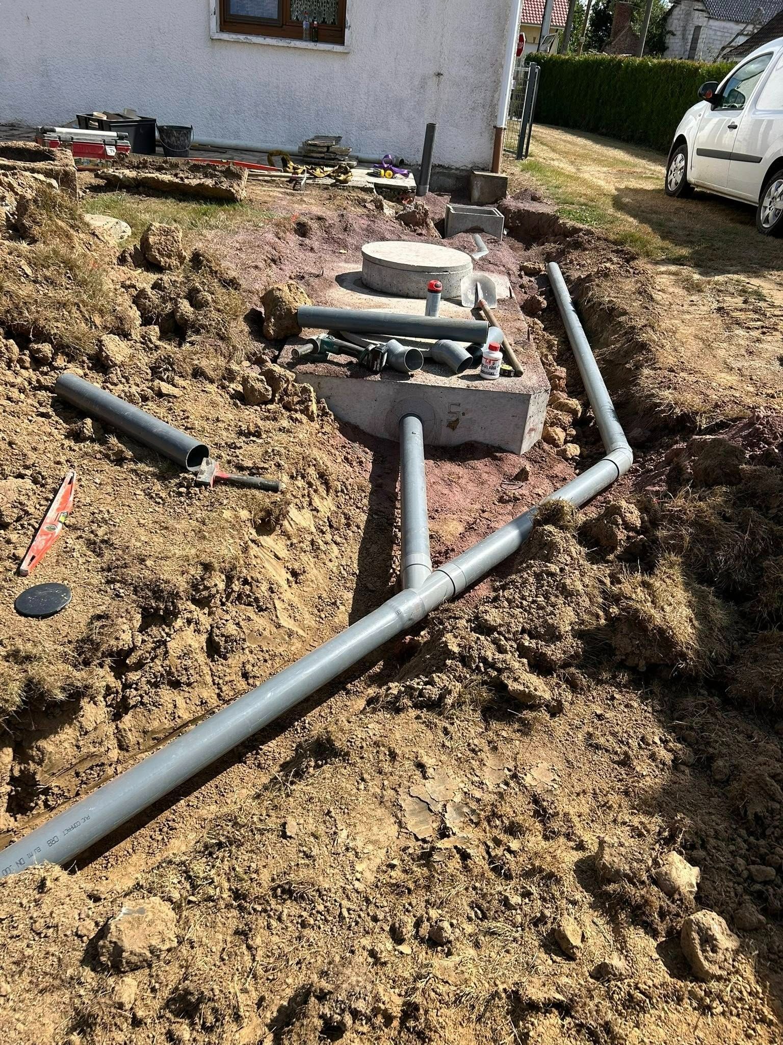 Chantier de construction avec des tuyaux en PVC gris dans une tranchée, près d'une maison et d'un véhicule.