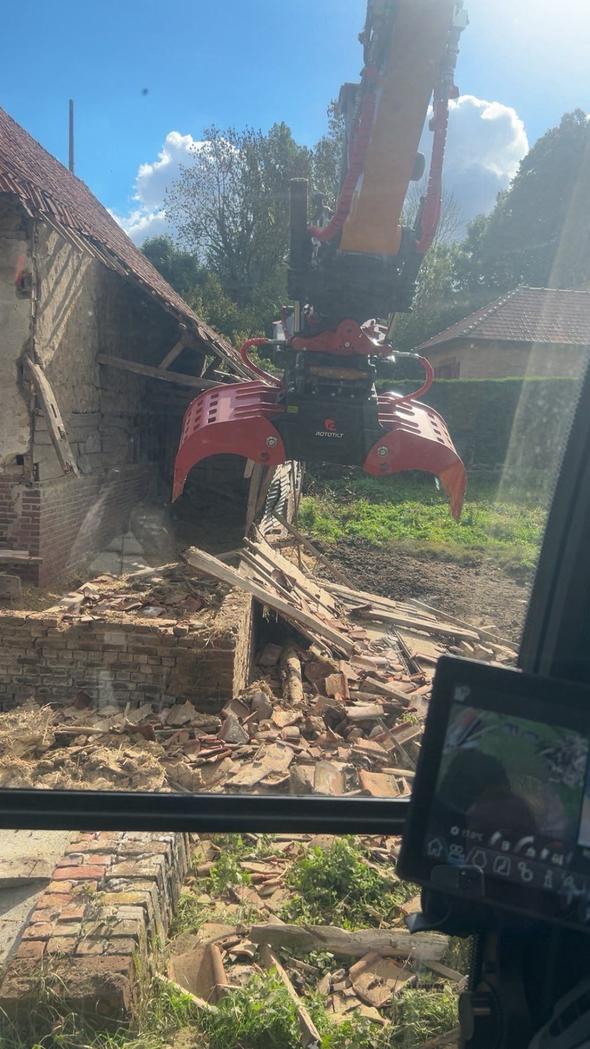 Une pelleteuse rouge démolit un bâtiment en briques, laissant apparaître des décombres sous un ciel bleu.