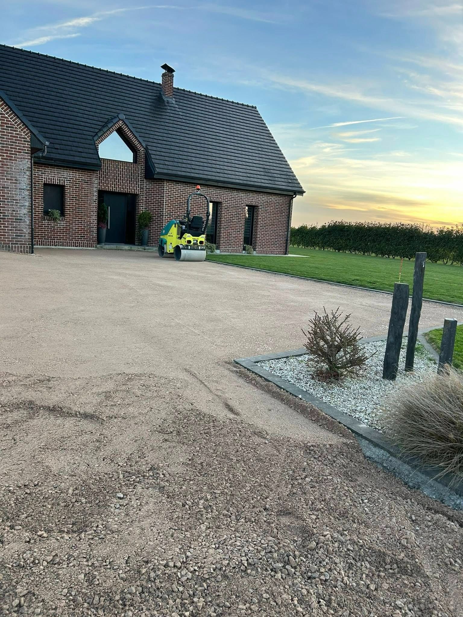 Une maison avec une allée en gravier ; un rouleau compresseur compacte le gravier. Coucher de soleil, ciel bleu.