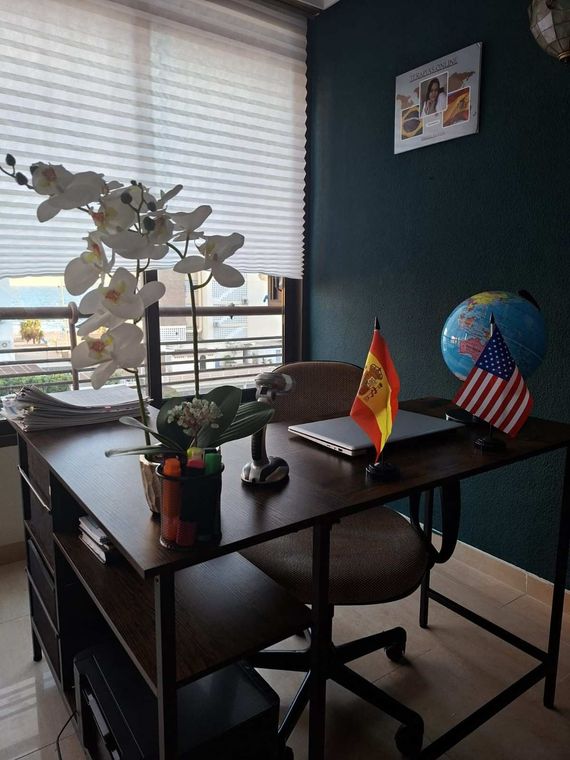 Un profesional sonriente, vestido con una camisa oscura y una bufanda estampada, está sentado en una oficina con libros y certificados al fondo.