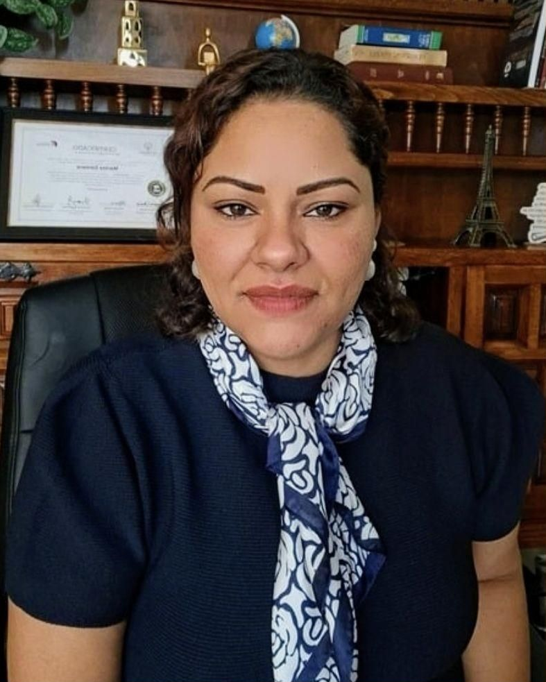 Una persona de cabello oscuro y bufanda azul estampada está sentada en una oficina con estanterías llenas de libros y objetos decorativos detrás.