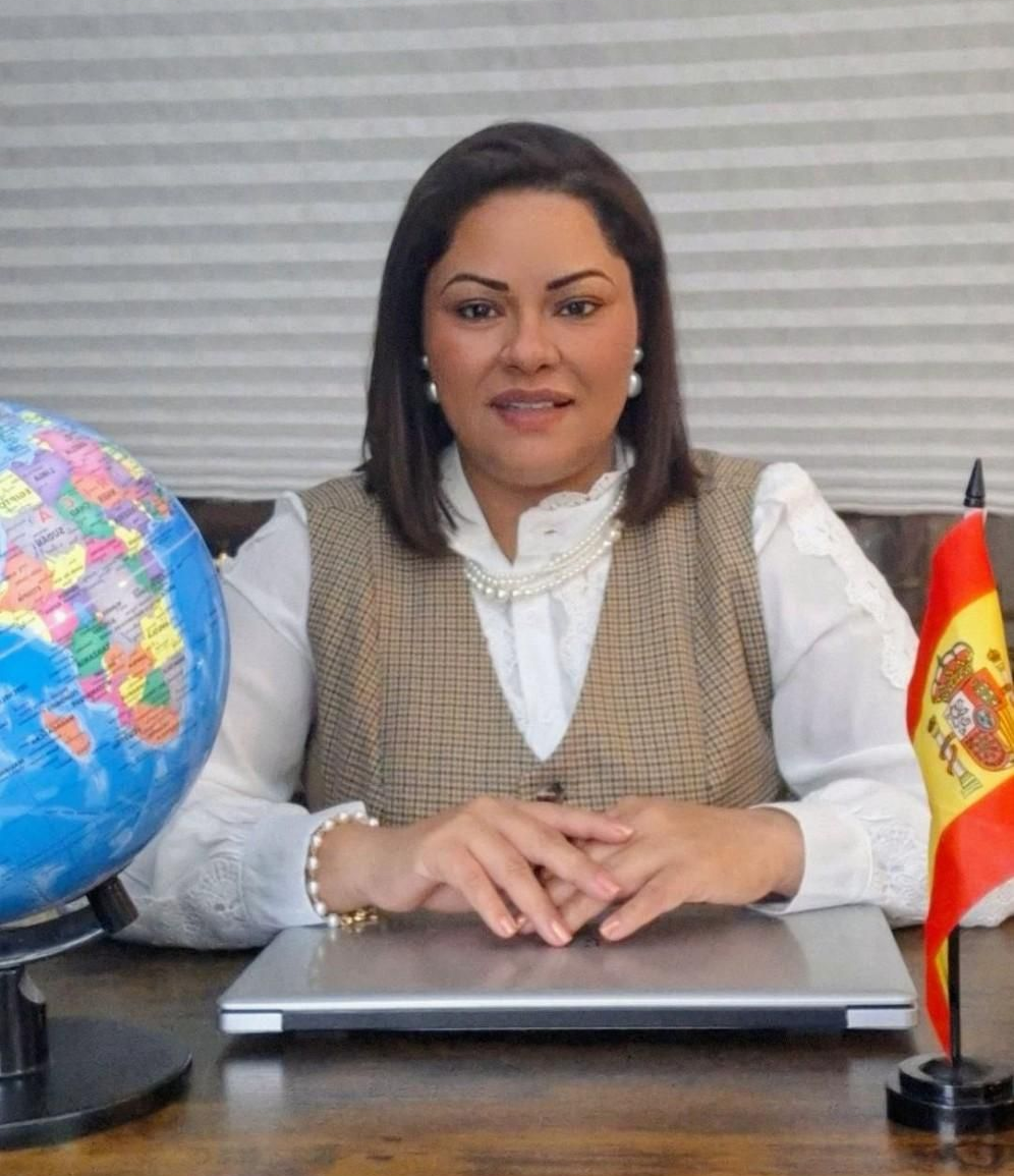 Una persona de cabello oscuro está sentada en un escritorio con una computadora portátil, un globo terráqueo y una pequeña bandera de España en un entorno profesional.