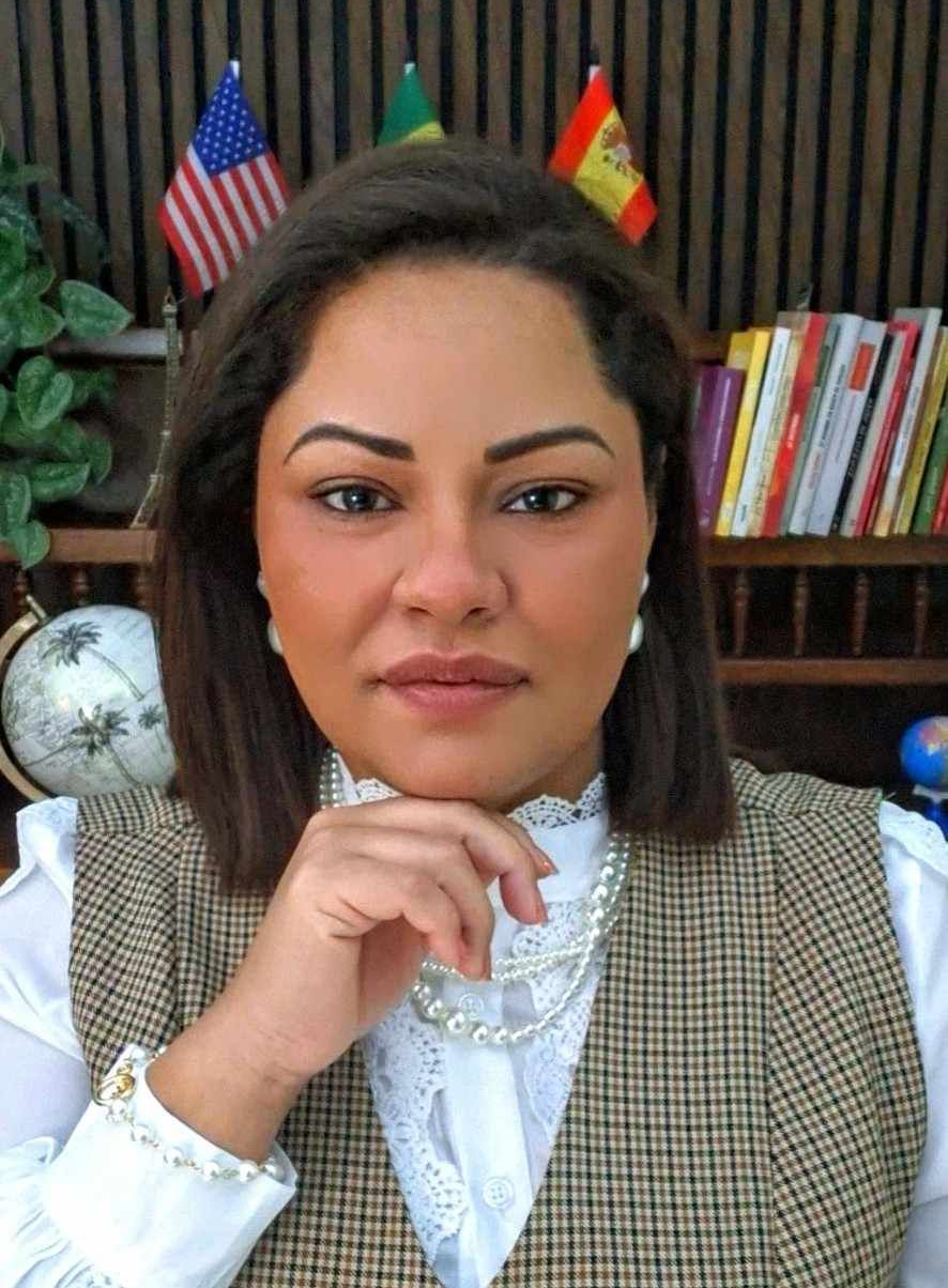 Una mujer profesional y sonriente, vestida con un blazer beige, está sentada frente a un escritorio con pequeñas banderas y una maqueta de la Torre Eiffel.