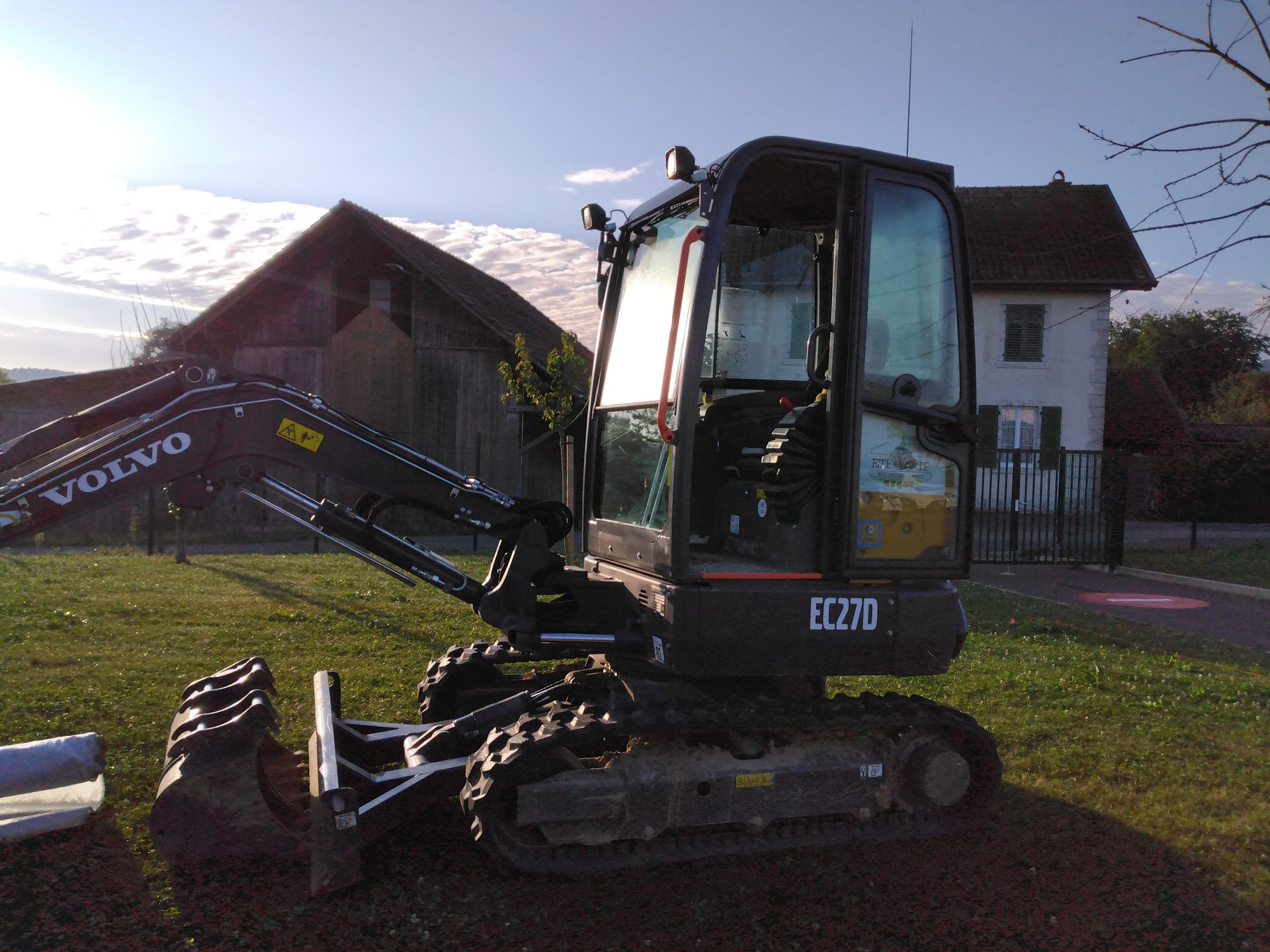 Une pelle mécanique Volvo EC270 est garée sur l'herbe, avec des bâtiments et le ciel en arrière-plan.