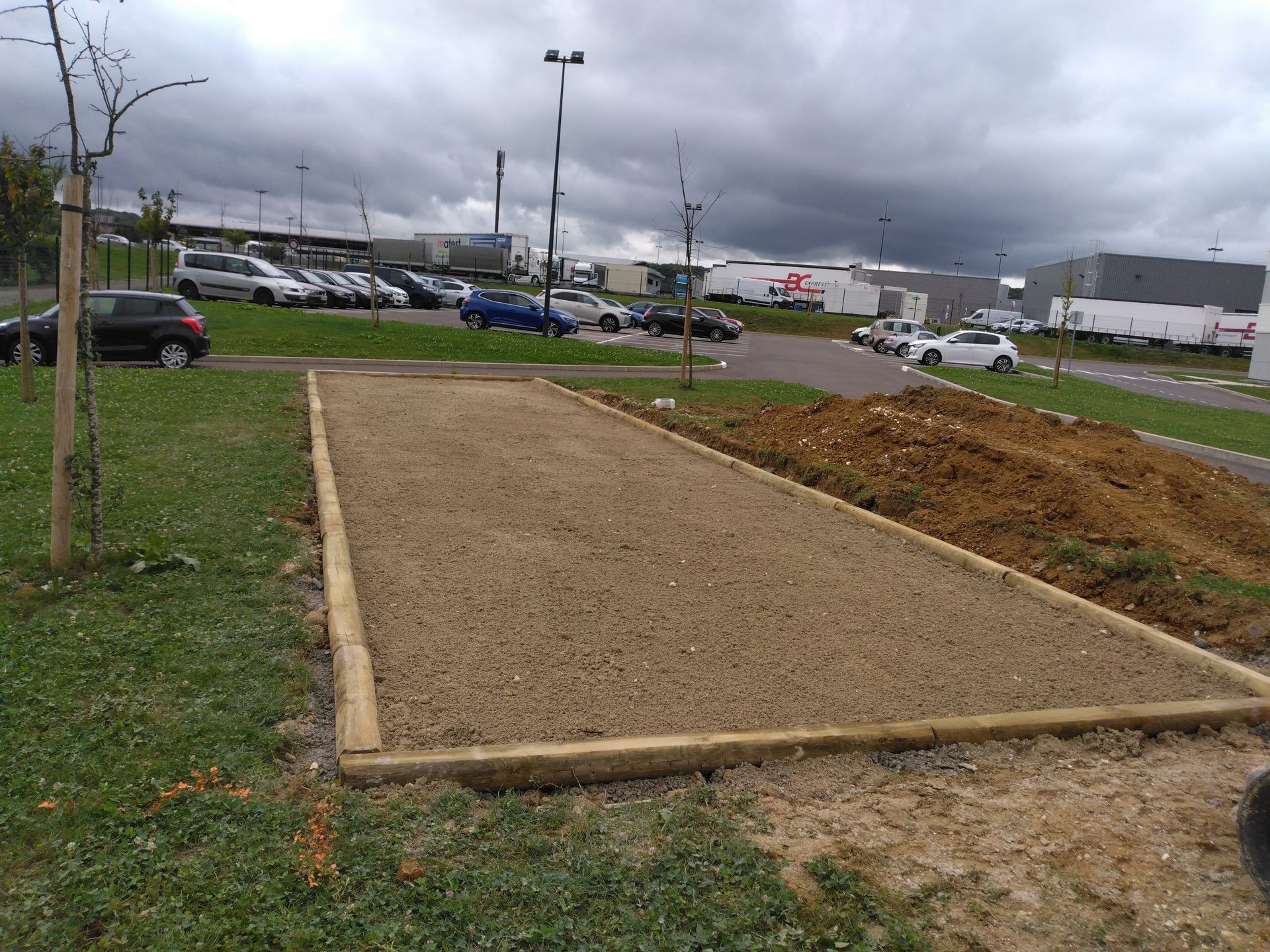 Terrain de pétanque en gravier bordé de bois, avec des voitures garées et un ciel nuageux en arrière-plan.