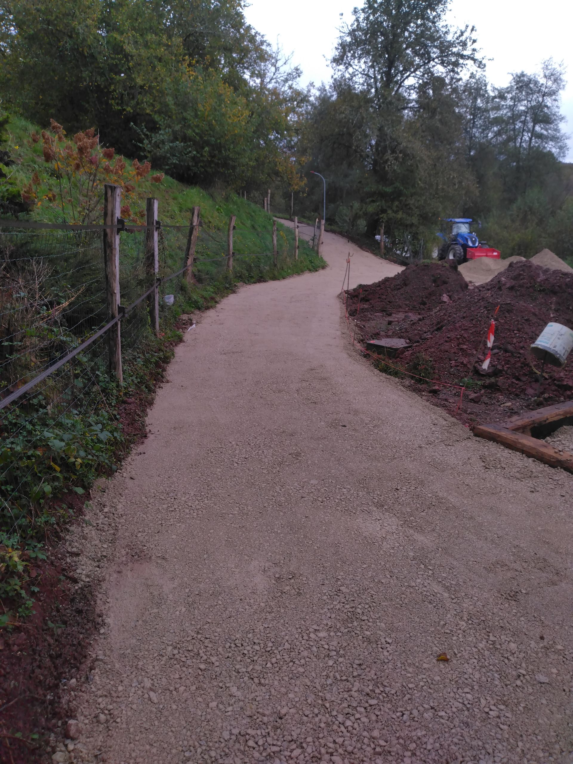 Chemin de gravier serpentant en montée, bordé d'une clôture et d'un talus herbeux. Engins de chantier visibles au sommet.