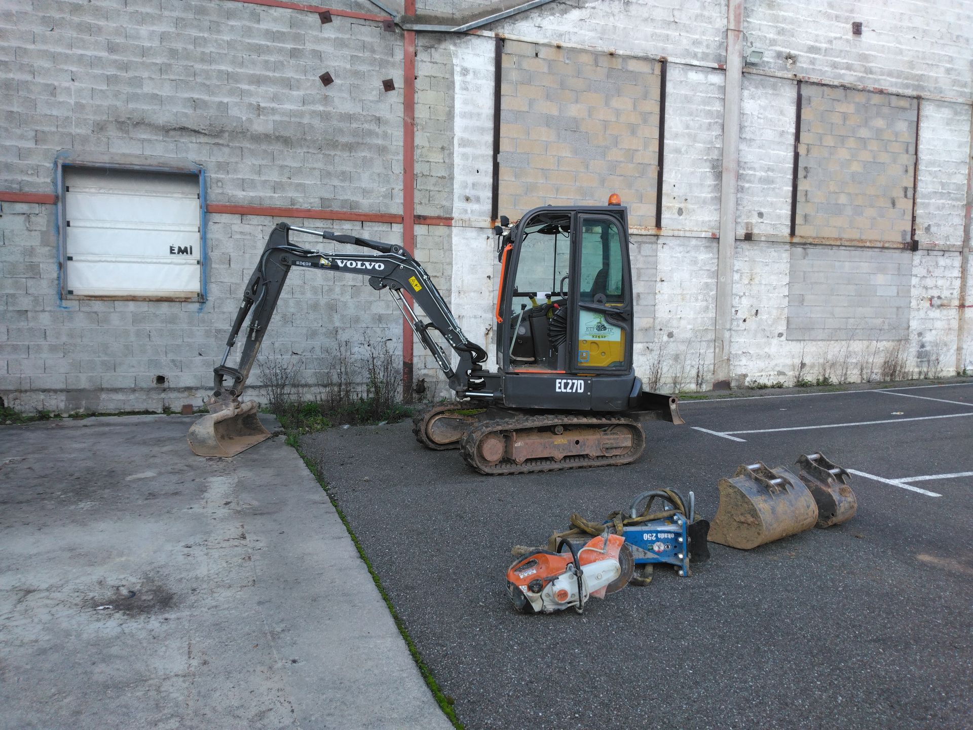 Une mini-pelle et des outils sont stationnés près d'un quai de chargement d'un bâtiment en briques.