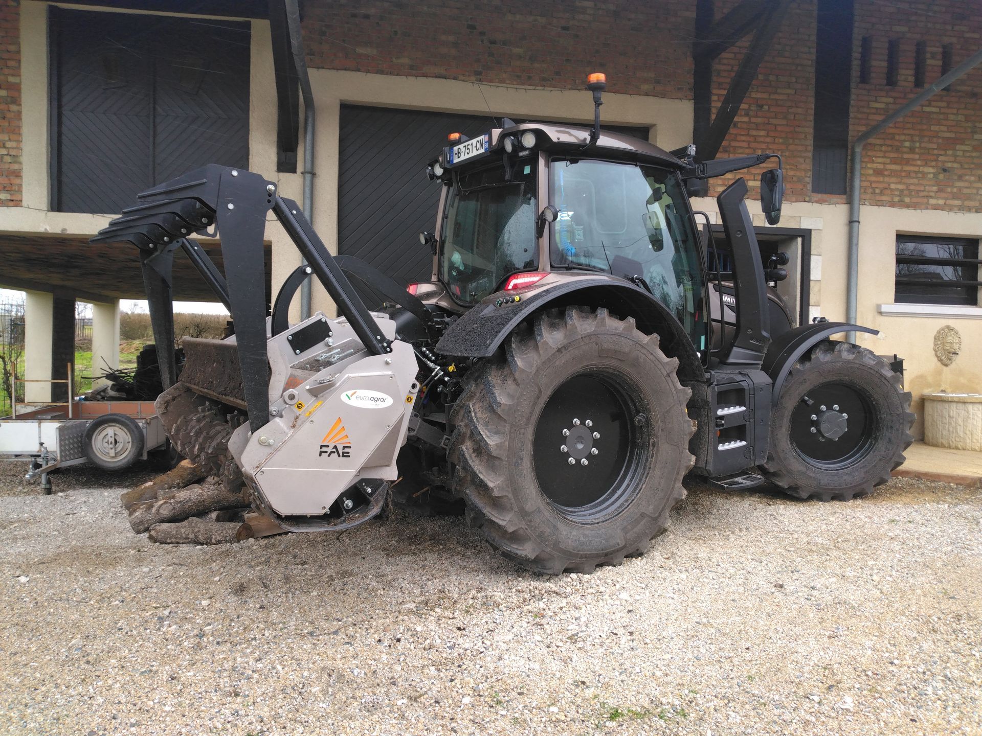 Tracteur noir équipé d'une débroussailleuse, stationné près d'un bâtiment sur un terrain gravillonné.