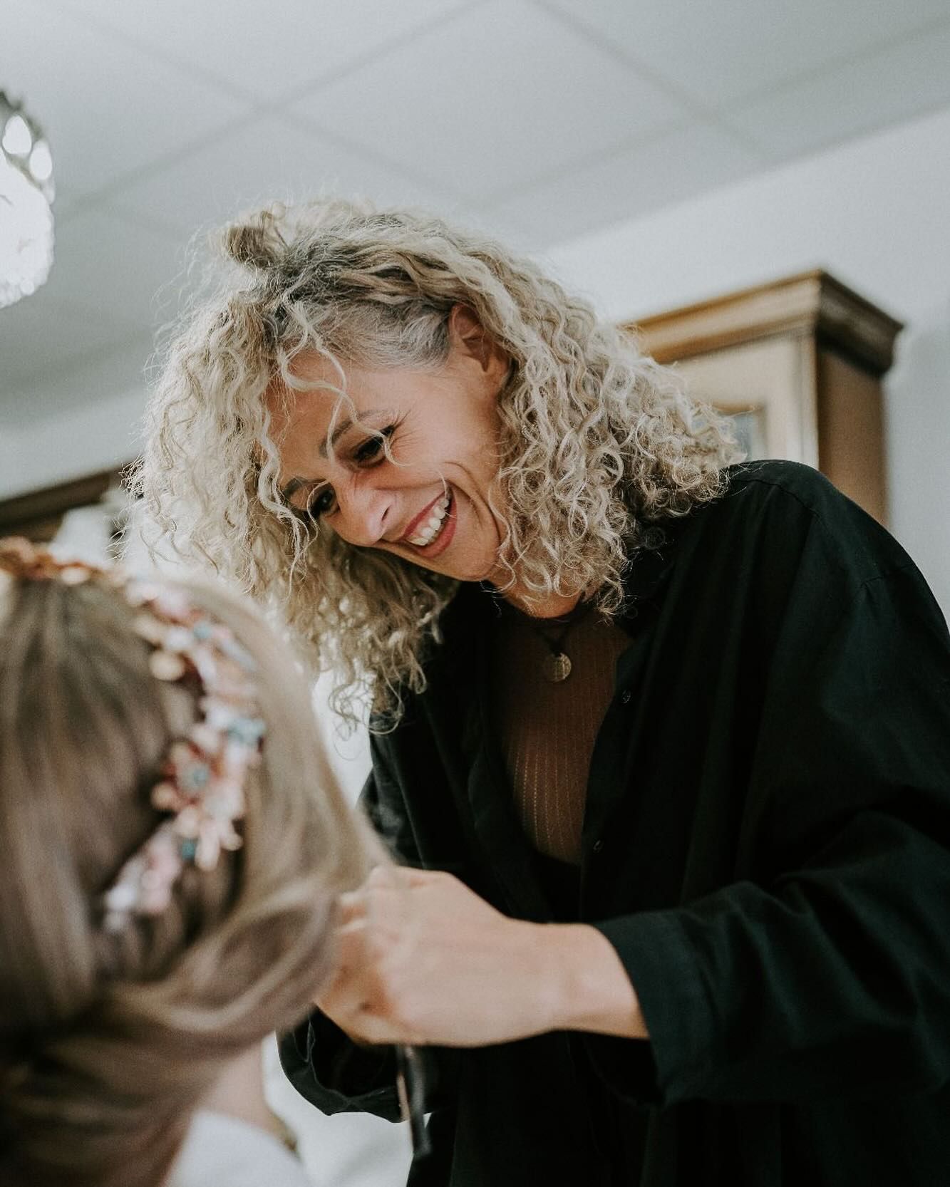 Una mujer con cabello rizado sonríe mientras peina el cabello de otra persona, en una habitación iluminada.