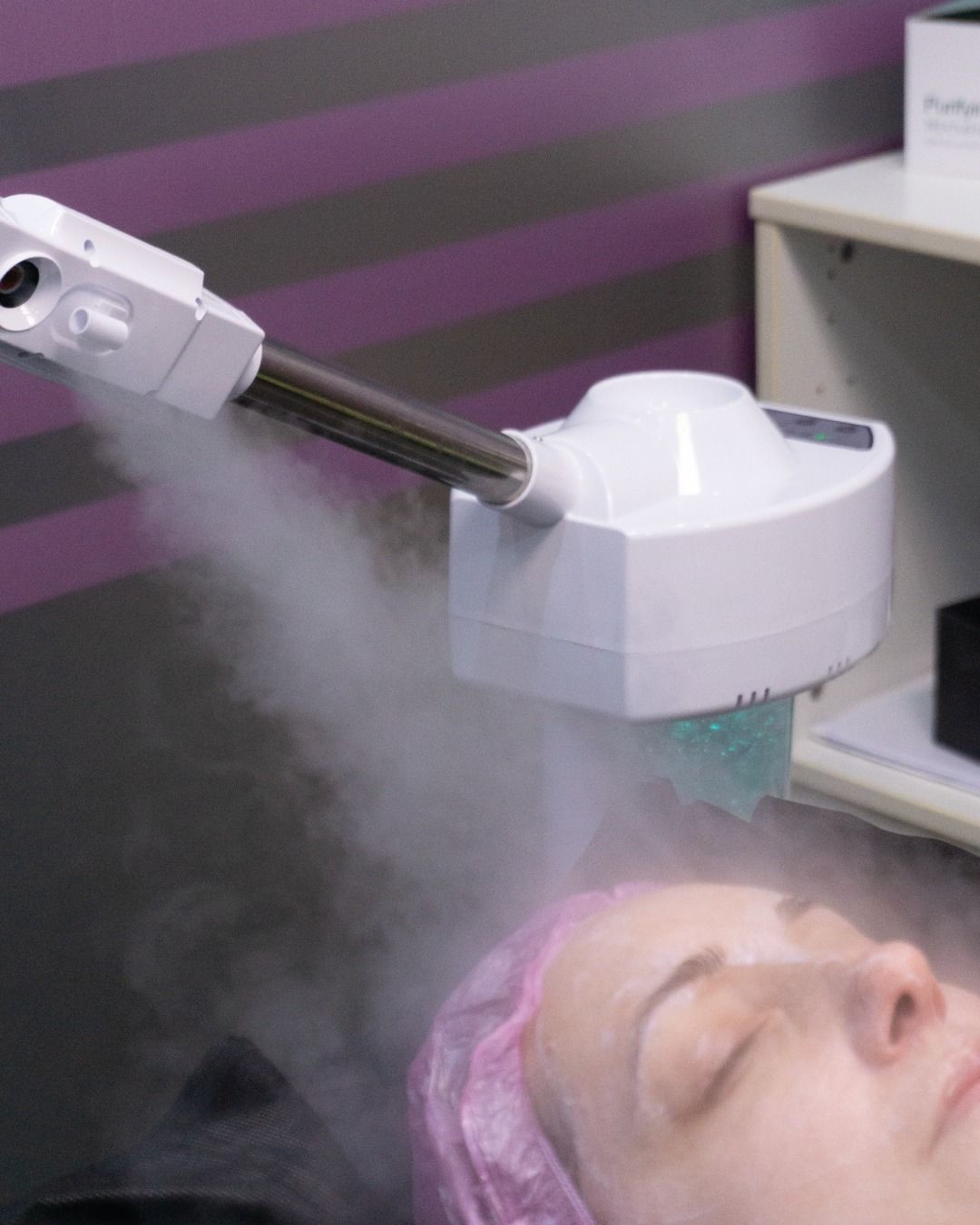 Vaporizador facial sobre el rostro de una persona en un spa; persona con gorra rosa, ojos cerrados, vapor visible.