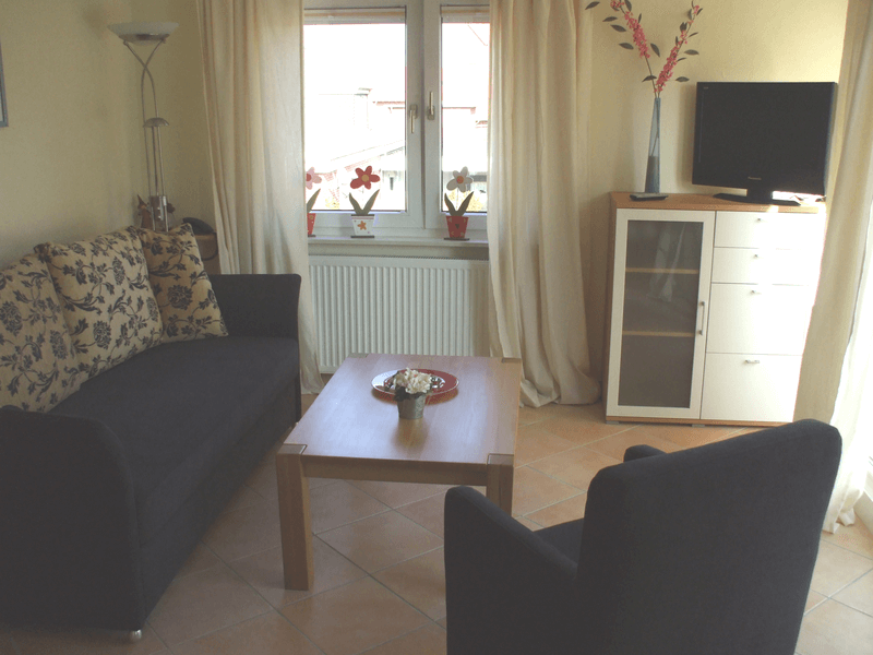 Wohnzimmer mit dunkelblauem Sofa, Sessel und Couchtisch. Weißer Schrank mit Fernseher, Fenster mit Vorhängen.