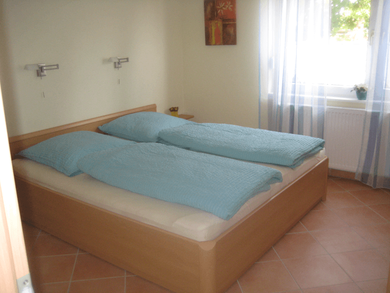 Schlafzimmer mit Doppelbett, hellblauer Bettwäsche, hellbraunem Holzrahmen, kleinem Fenster und Fliesenboden.