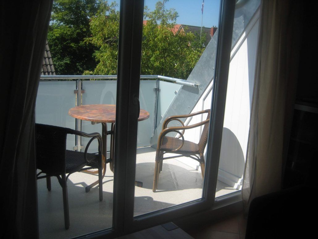 Balkon mit Tisch und zwei Stühlen, durch Glastüren zu sehen; draußen sind Bäume und blauer Himmel erkennbar.