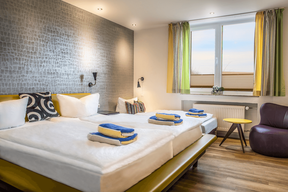 Hotelzimmer mit zwei Einzelbetten, dekorativer Wand, Fenster mit Vorhängen und einem kleinen Stuhl.