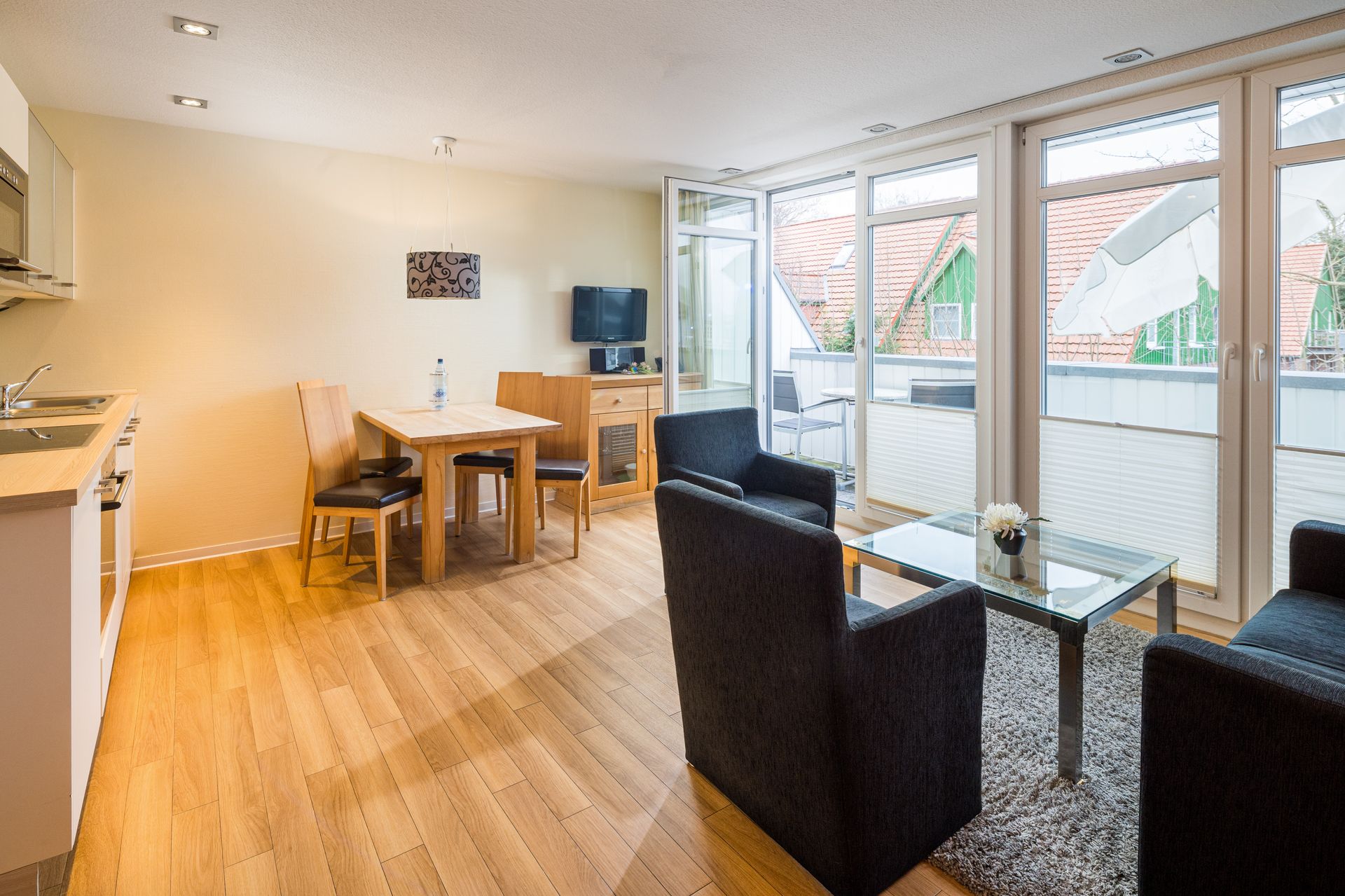 Wohnzimmer mit Küche, Essbereich und Zugang zum Balkon. Helle Holzböden, schwarze Stühle.