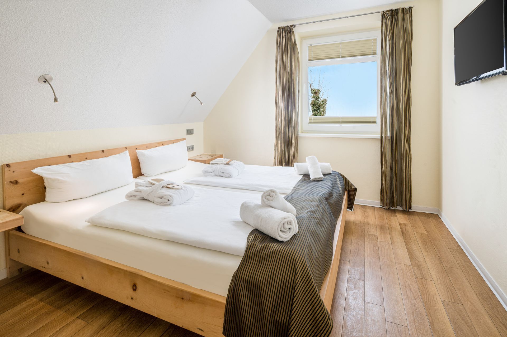 Schlafzimmer mit zwei Betten, weißer Bettwäsche und Holzrahmen. Fenster mit Vorhängen, Holzboden und Wandfernseher.