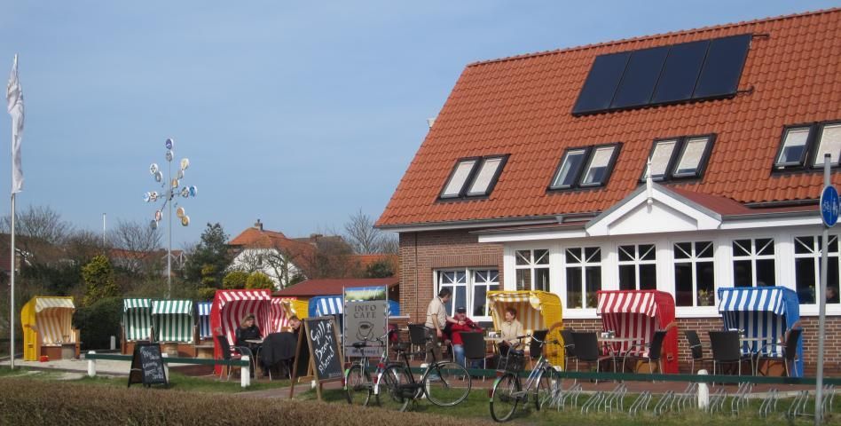 Café mit Strandstühlen, Fahrrädern und Solarpaneelen auf einem roten Dach. Leute auf der Terrasse.