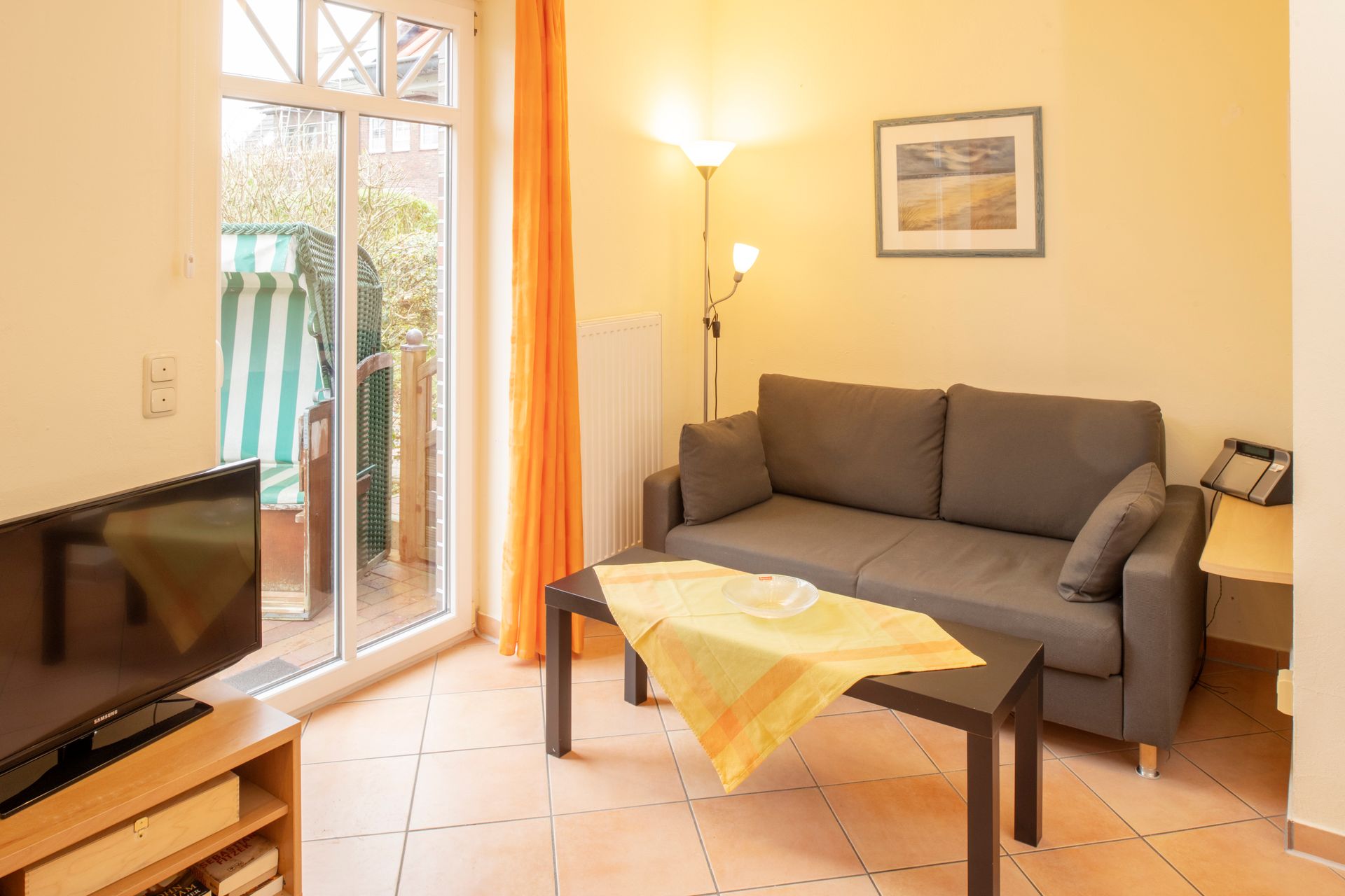 Wohnzimmer mit grauem Sofa, Fernseher, Glastür zum Balkon und einem Couchtisch mit gelber Tischdecke.