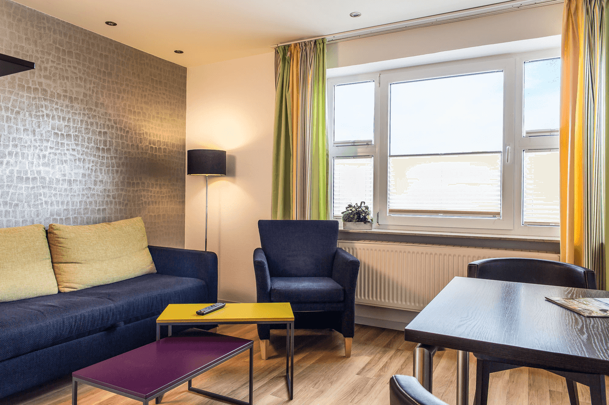 Wohnzimmer mit blauem Sofa, Sessel und Tisch, mit gemusterter Tapete, Fenster und farbenfrohen Vorhängen.