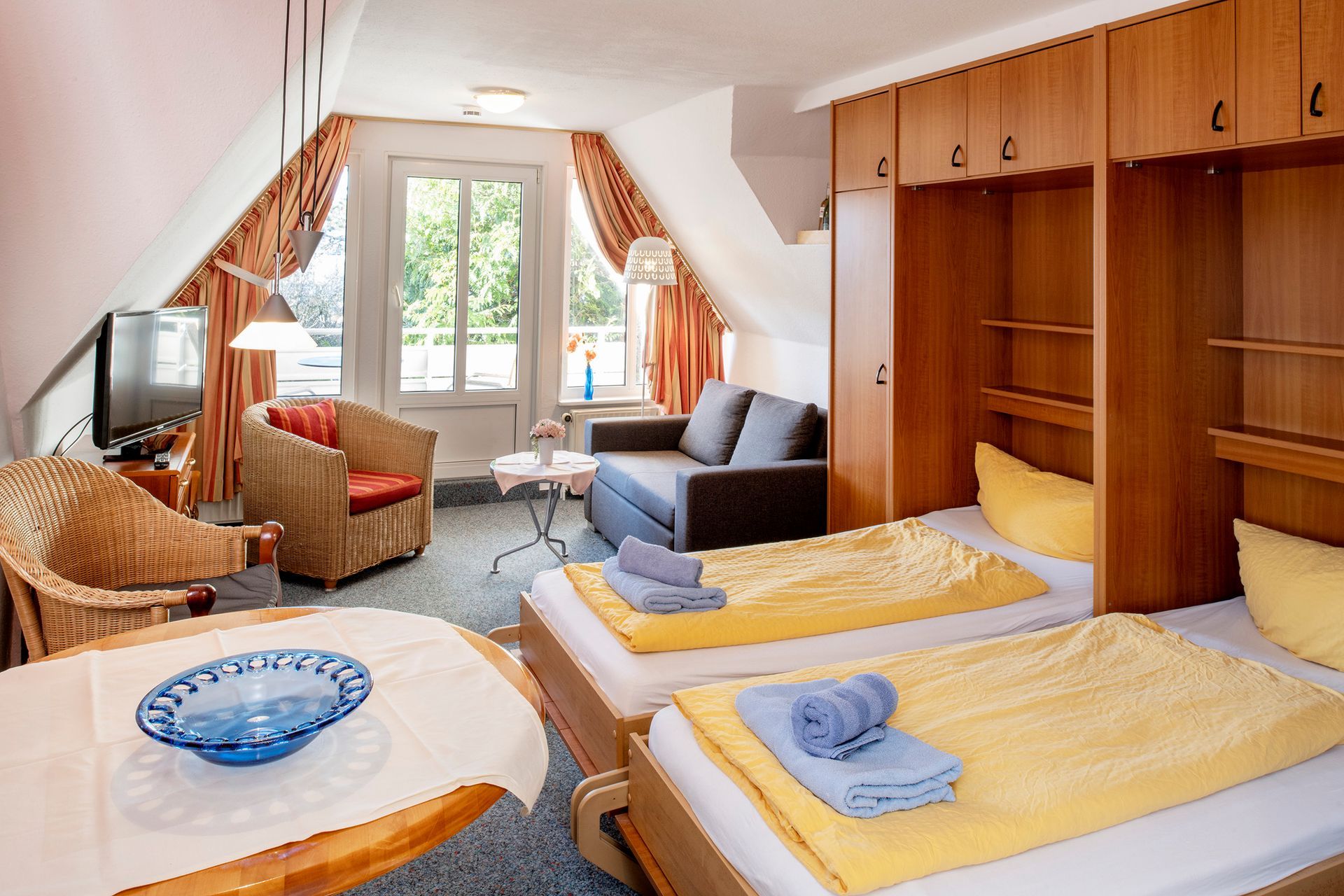 Hotelzimmer mit zwei Betten, Sitzecke, Fernseher und Zugang zum Balkon. Eingebauter Holzschrank.