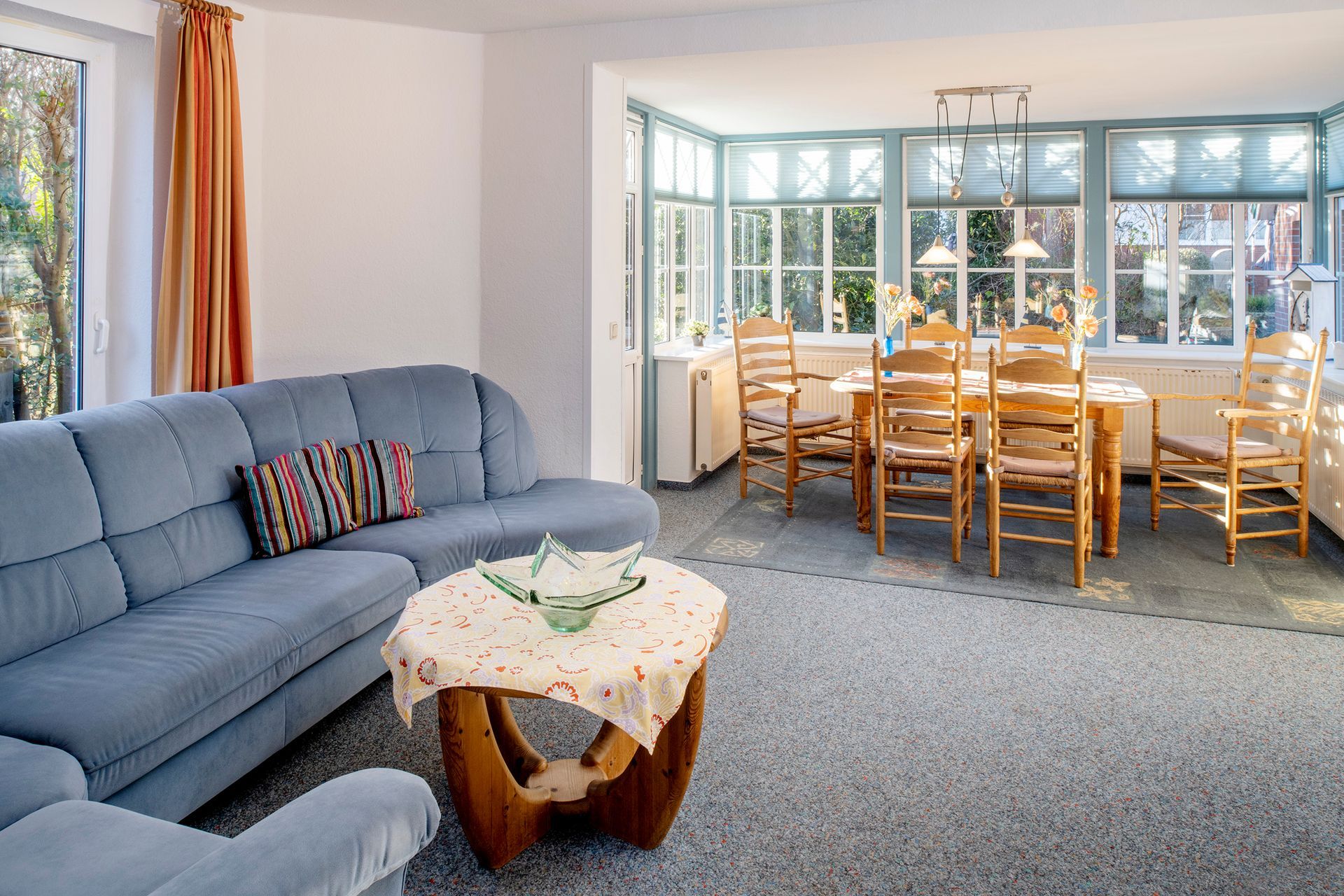 Wohnzimmer mit blauem Sofa, Holztisch und Stühlen sowie großen Fenstern.