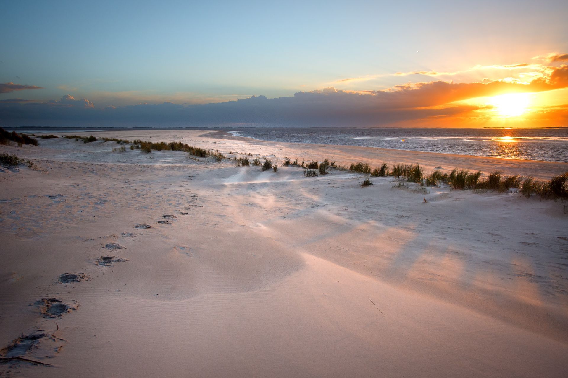 Sonnenuntergang über einem Strand mit Fußspuren im Sand und wehender Wind.