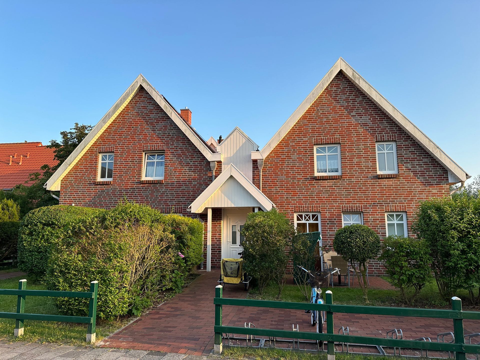 Backsteinhaus mit zwei Giebelteilen, weißen Zierelementen, grüner Hecke und Zaun, klarer blauer Himmel.