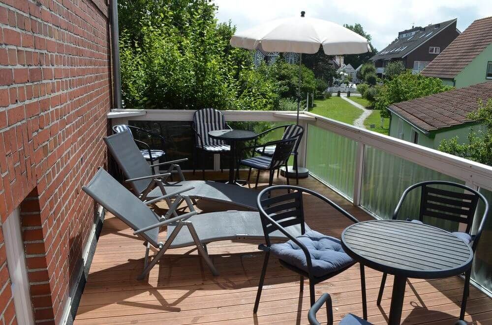 Balkon mit Liegestühlen, Tischen und einem Sonnenschirm. Links eine Backsteinmauer, im Hintergrund Bäume und Gebäude.