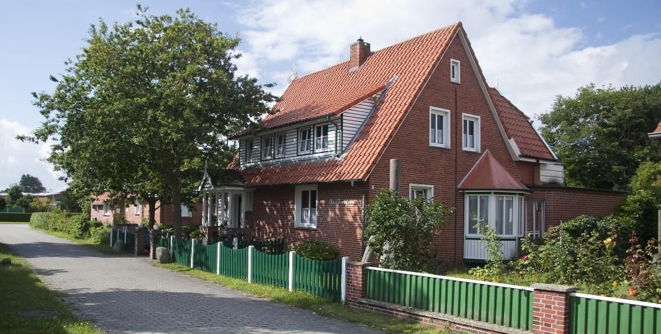 Backsteinhaus mit rotem Dach, grünem Zaun und kleinen Bäumen an einem sonnigen Tag.